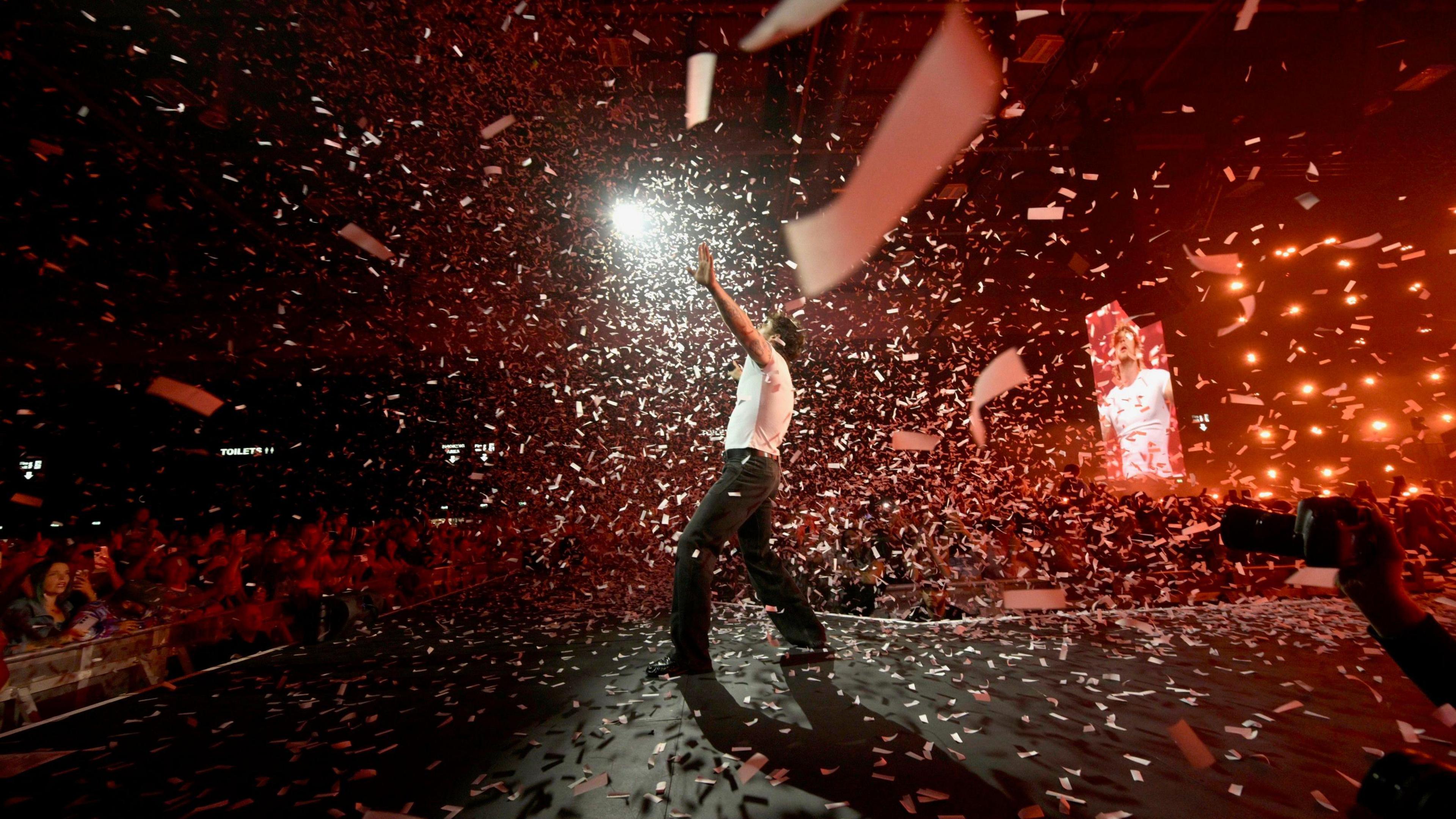 A side view of singer Tom Grennan on stage with the audience in front. He is wearing a white T-shirt and black trousers and there is confetti falling from the ceiling around him.