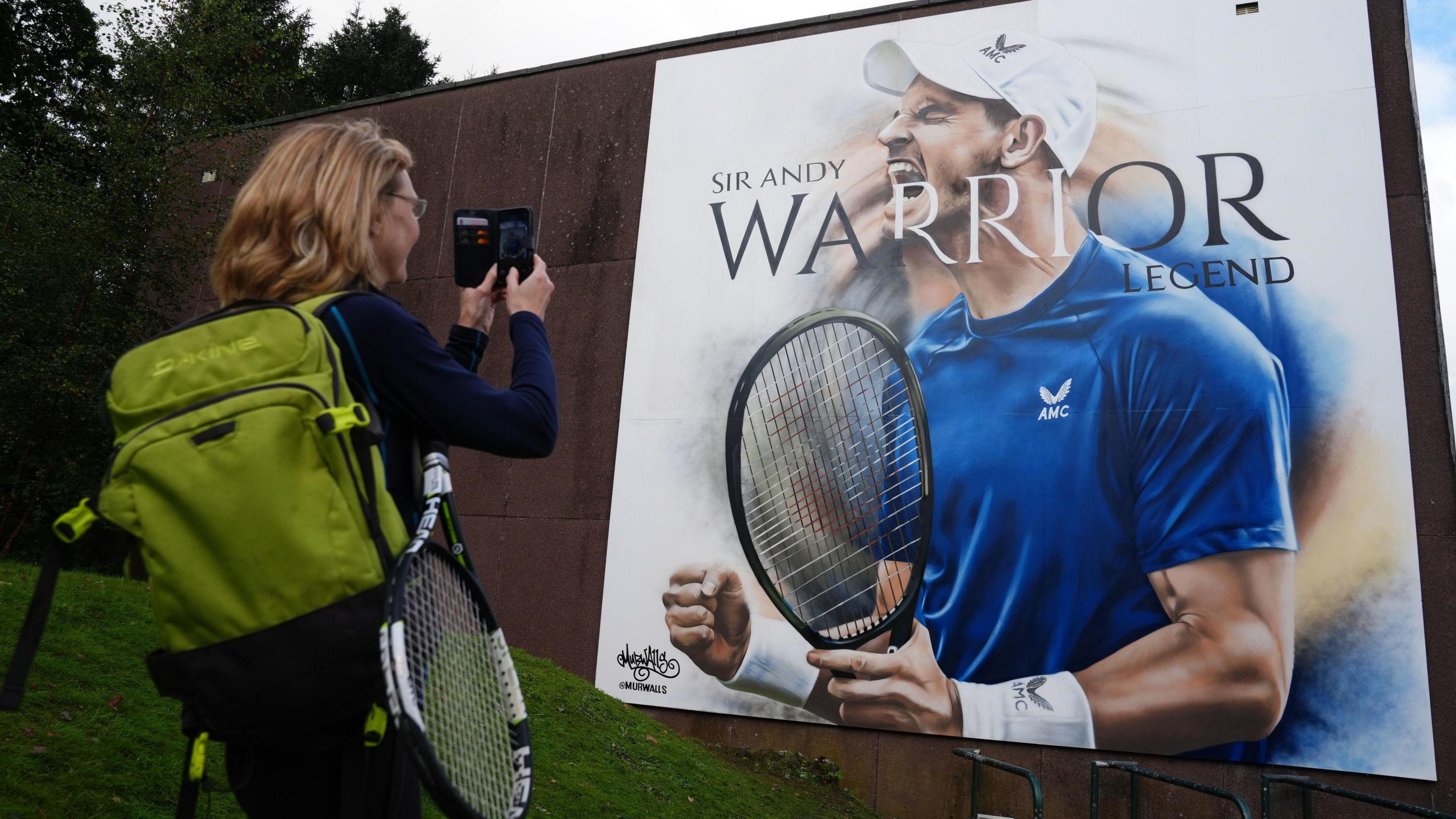 Woman carrying a green backpack and a tennis racket uses her phone to take a photograph of a large artwork of Andy Murray. It is a picture of Murray in a blue t-shire and white cap. It says Andy Murray. Warrior. Legend.