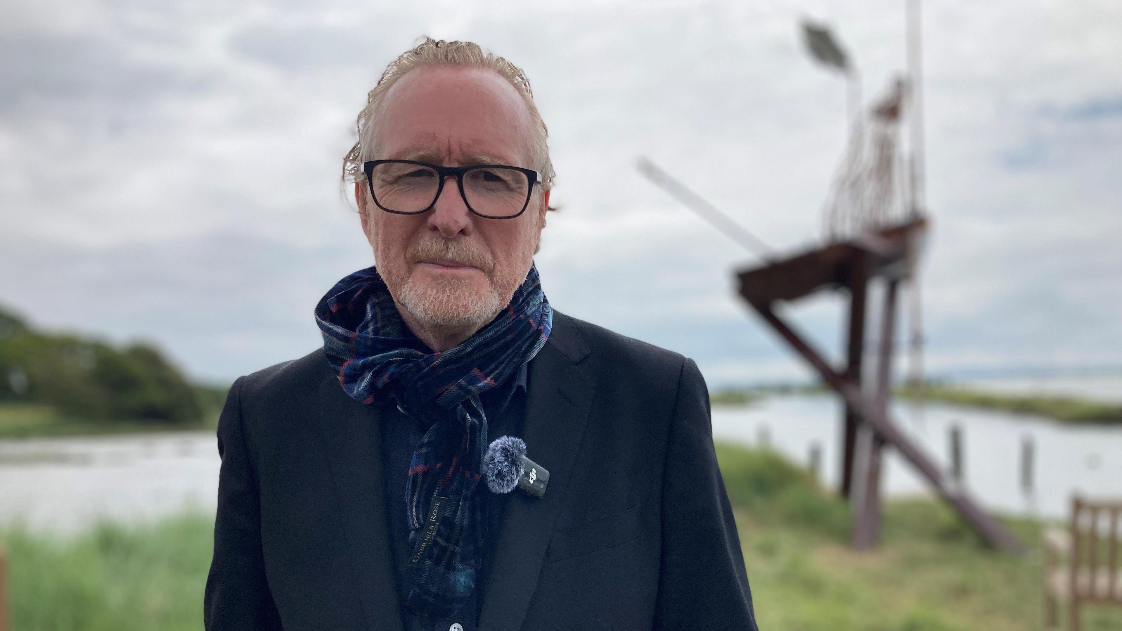 Alistair Morrison, a white man in his 50s, with grey/ginger hair slicked back and a short, full beard wearing black framed glasses, a blue and red tartan scarf and dark blue blazer looks at the camera, with the sculpture in the background