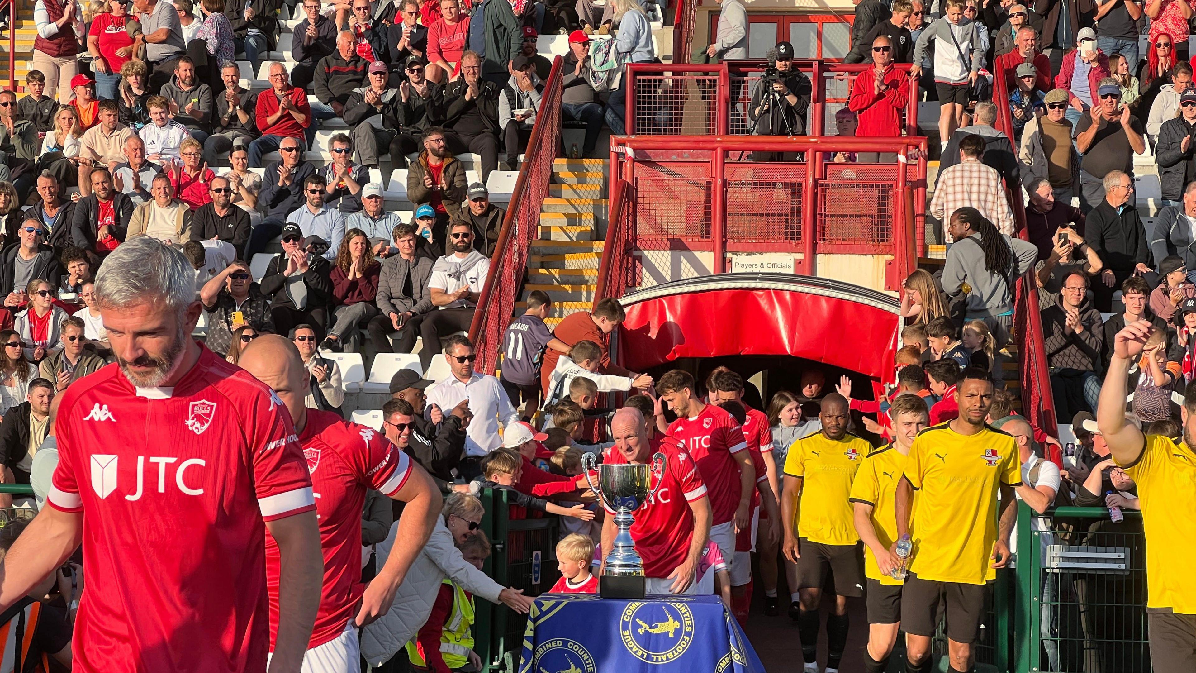 Players come out of the tunner and go past the trophy as fans in the background cheer on from the stand.