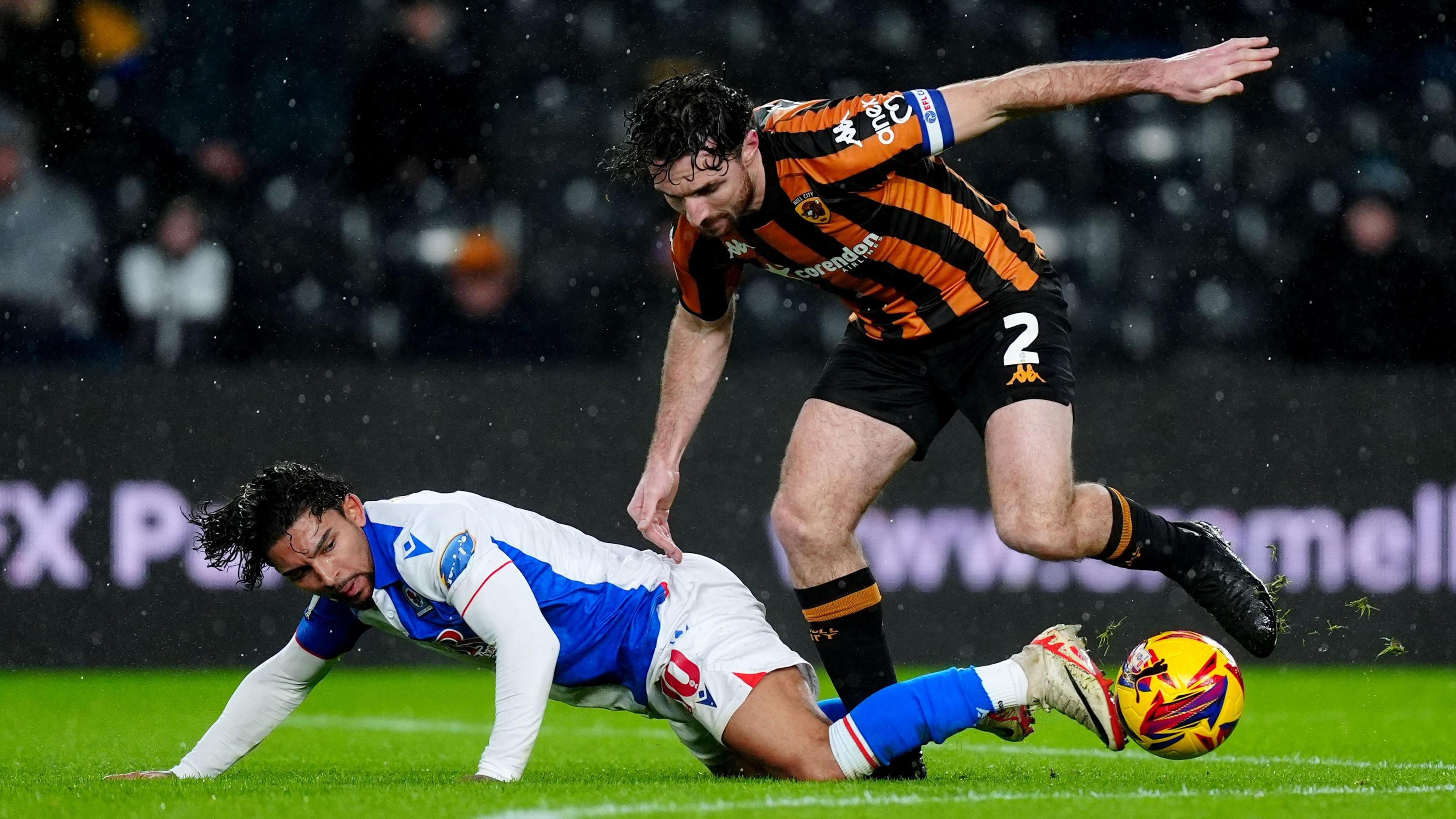 Hull City captain Lewie Coyle avoids a tackle during a match. He is on the right of the image and is wearing a black and amber shirt, black shorts, black and amber socks and a pair of black football boots. The yellow, red and blue football is up in the air after bouncing off the green grass pitch. Tyrhys Dolan, a Blackburn Rovers player, is laid on the floor and is wearing a blue and white shirt, white shorts and blue socks.