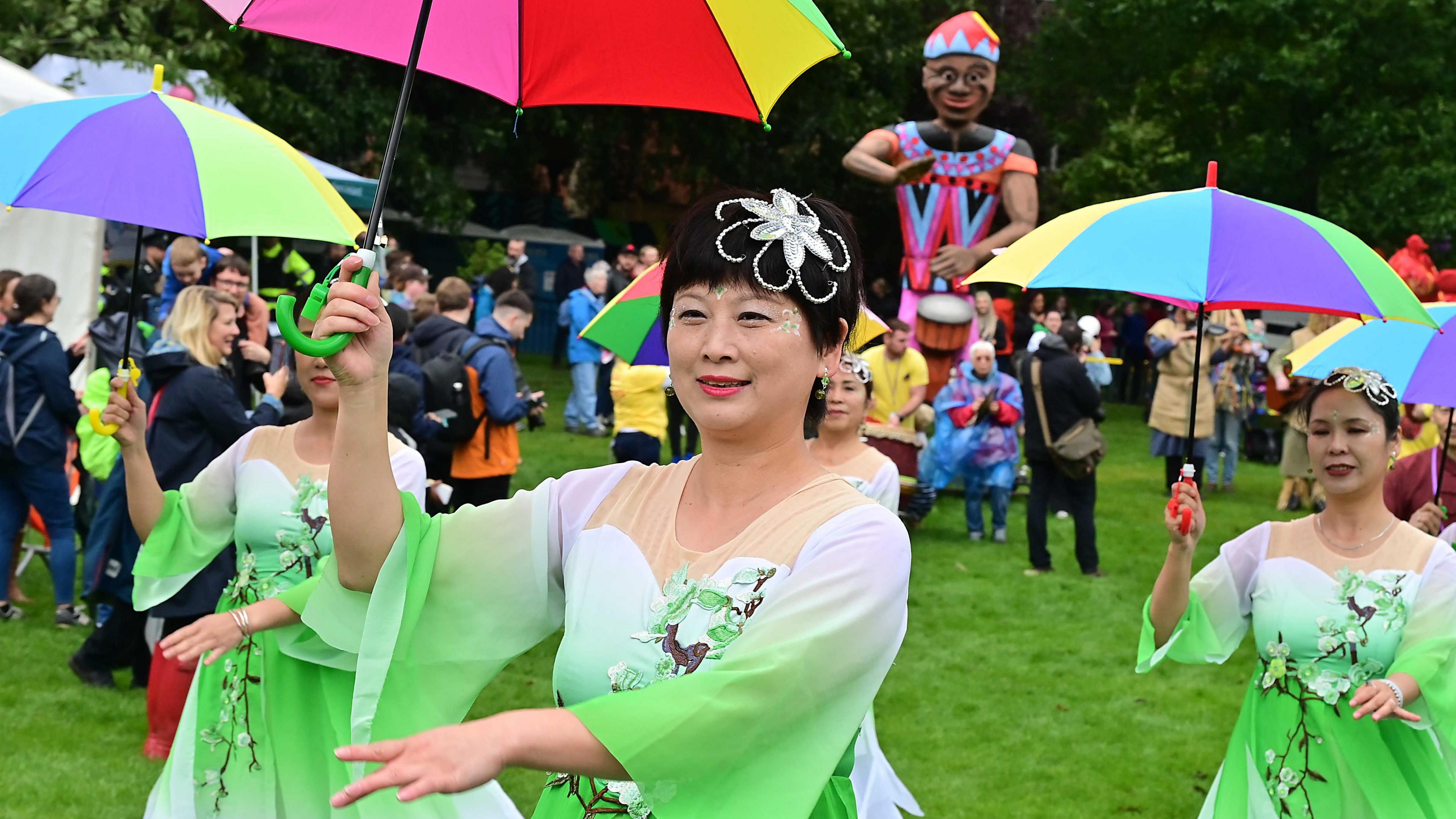 Women in green and white dresses walk along festival with rainbow umbrellas. 