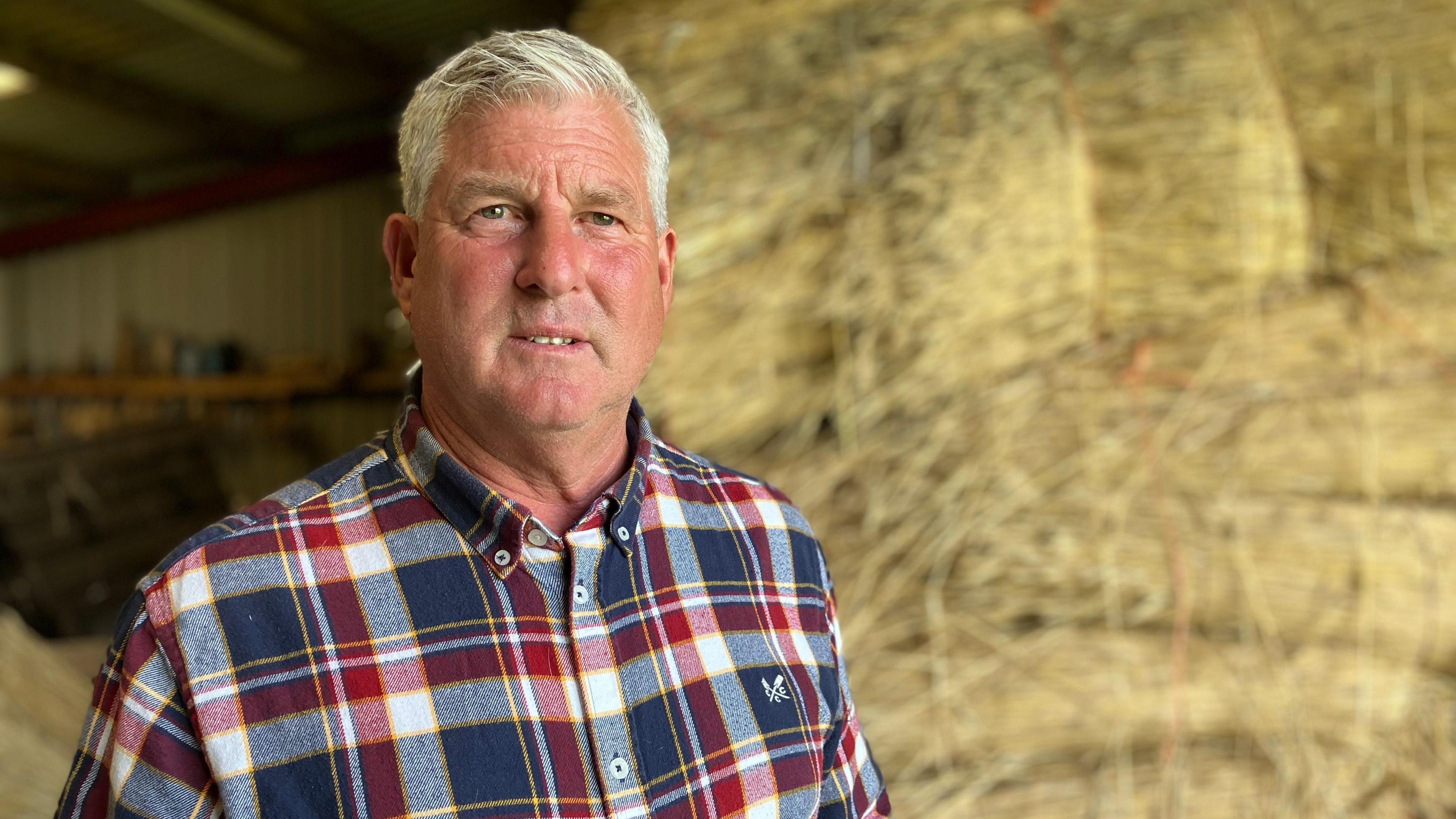 A man is looking at the camera. He has grey short hair and is wearing a blue and red checked shirt. He is stood in front of straw bales. 