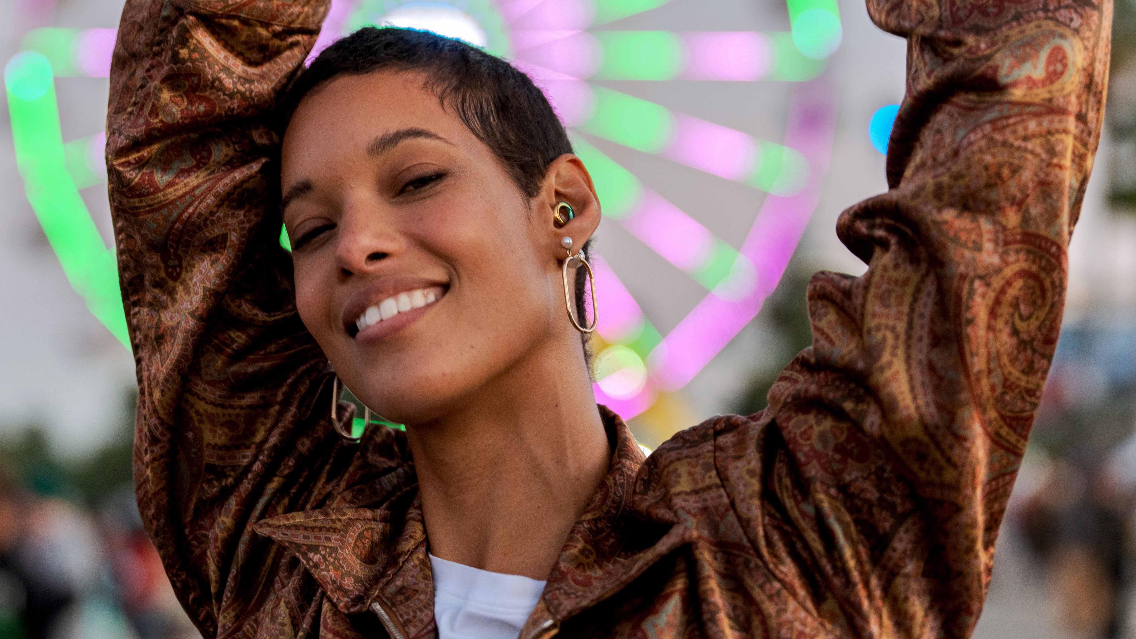 A young pistillate   dances astatine  dusk successful  beforehand   of a neon-lit Ferris instrumentality    astatine  a euphony  festival. As good   arsenic  ample  hoop earrings, she wears a rainbow-coloured ear-plug arsenic  extortion  for her hearing.