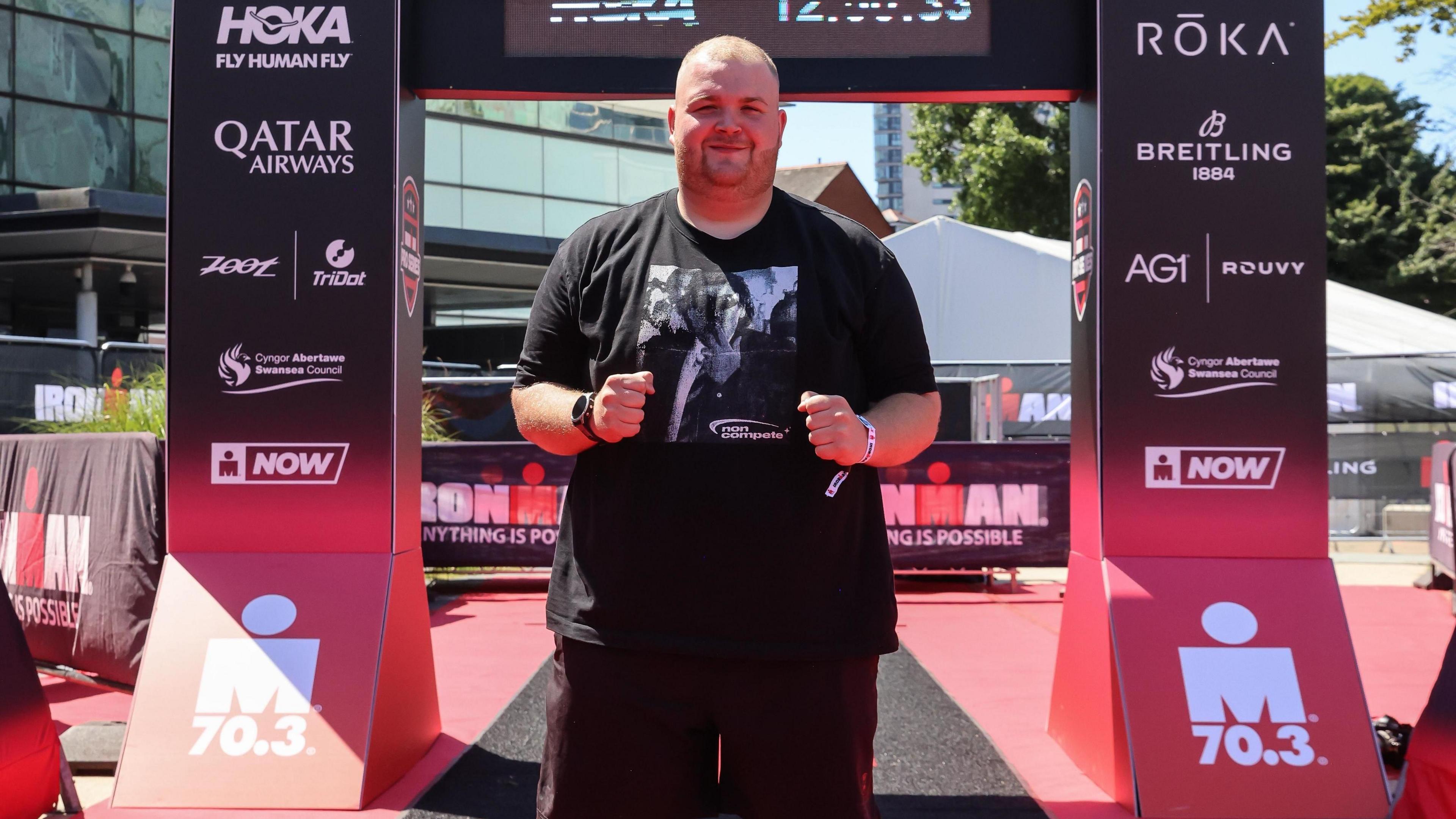 Owen Peddle at Ironman-branded arch in black t-shirt and shorts. He has a shaved head and short trimmed facial hair and smiles with his fists held out in front of his chest