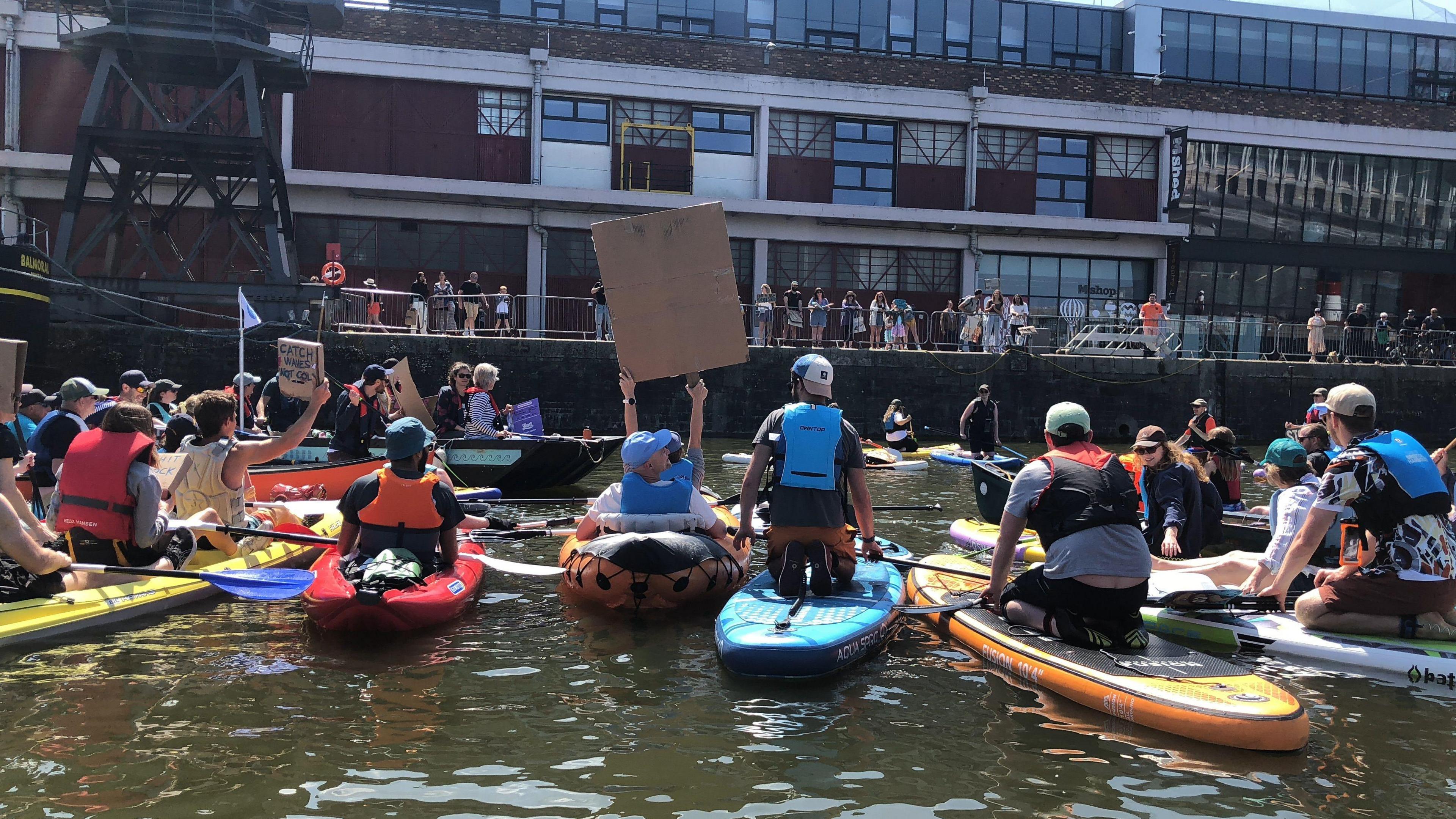 A group of people on paddleboards taken from the back. They are in a harbour holding signs to a crowd of people on the harbour side. They have multicoloured boards and life jackets.