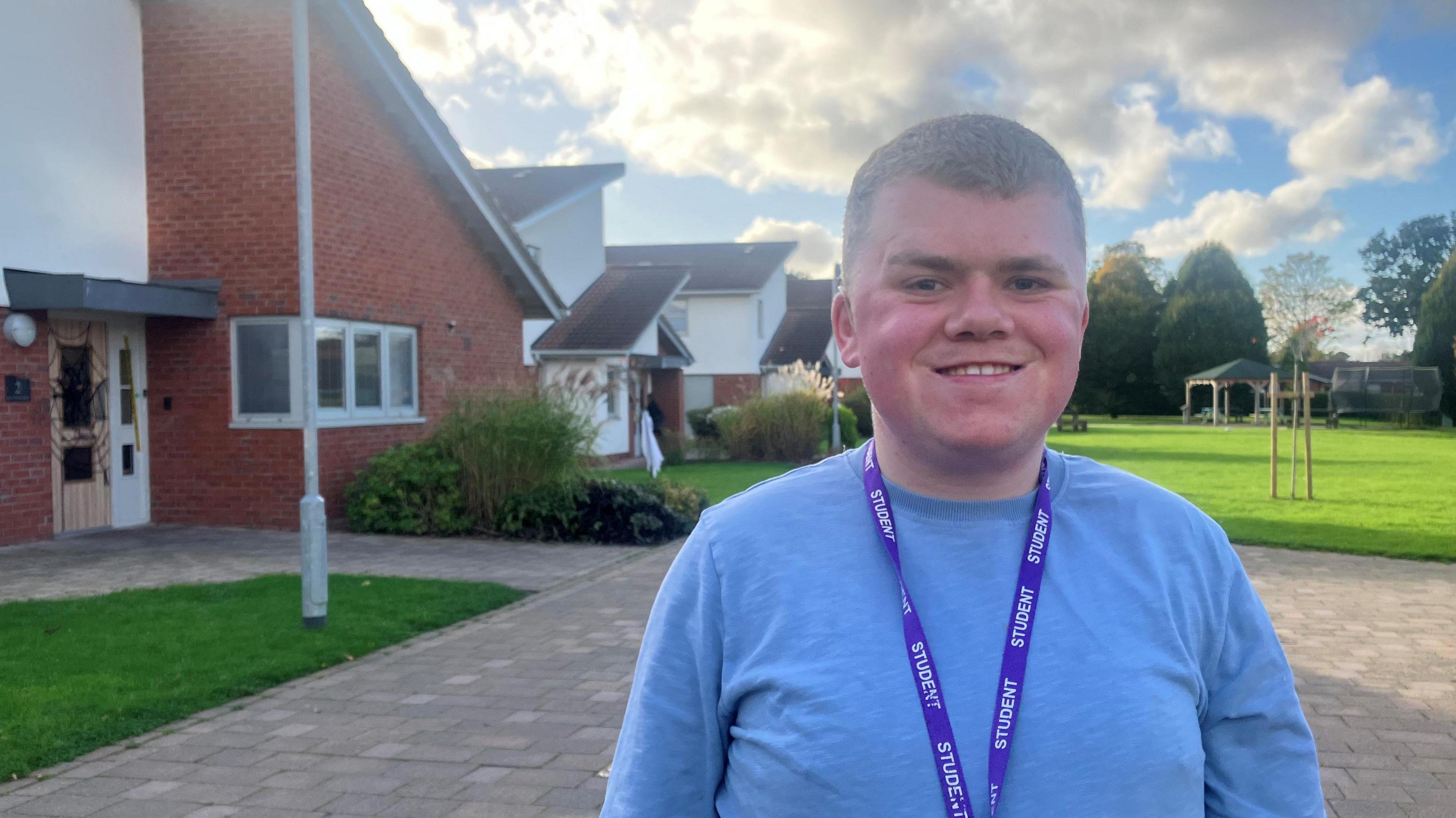 Photo of Max Pennells, a 20 year-old student at the Royal College Manchester. He is wearing a light blue shirt and a lanyard, and is standing in front of a lawn and a modern, brick-built building. 