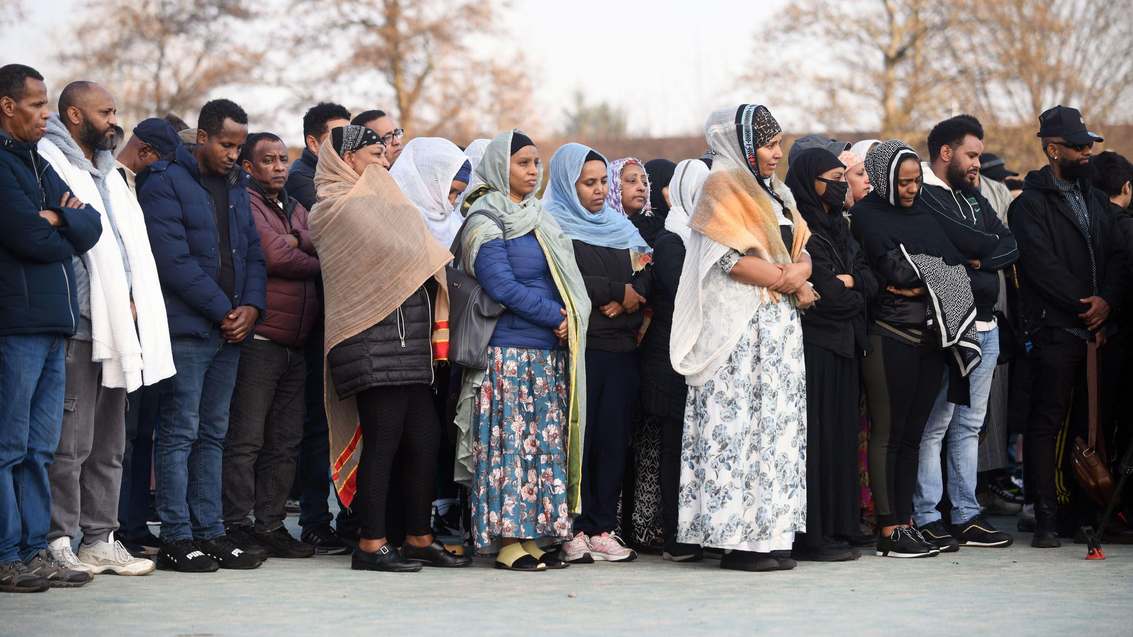 A row of sombre looking people, the women in head dresses, standing in the open air, clearly listening to someone speaking. Some have their heads dipped and their hands crossed. 