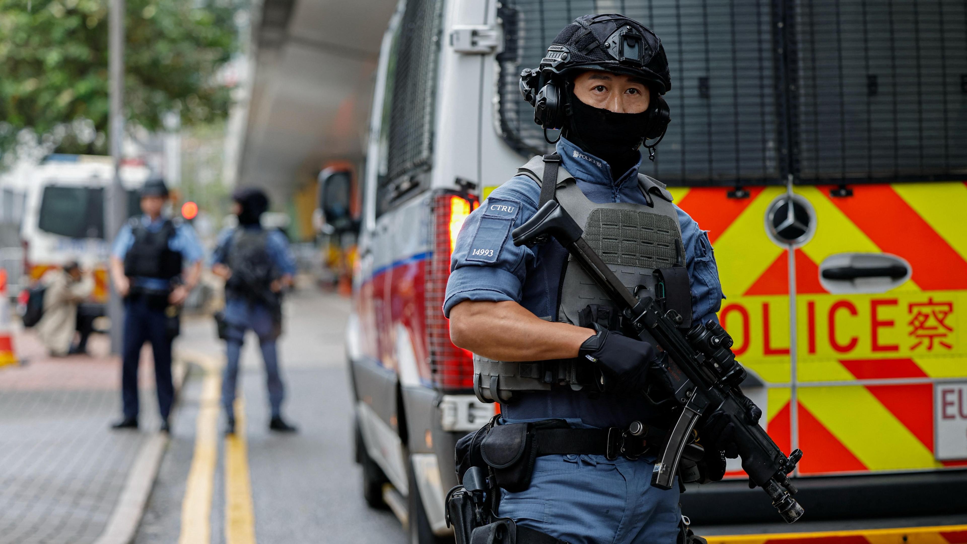 Armed police stand guard with weapons and wearing helmets in front of a police van on a street under a bridge as a prison van arrives at the West Kowloon Magistrates' Courts building, 