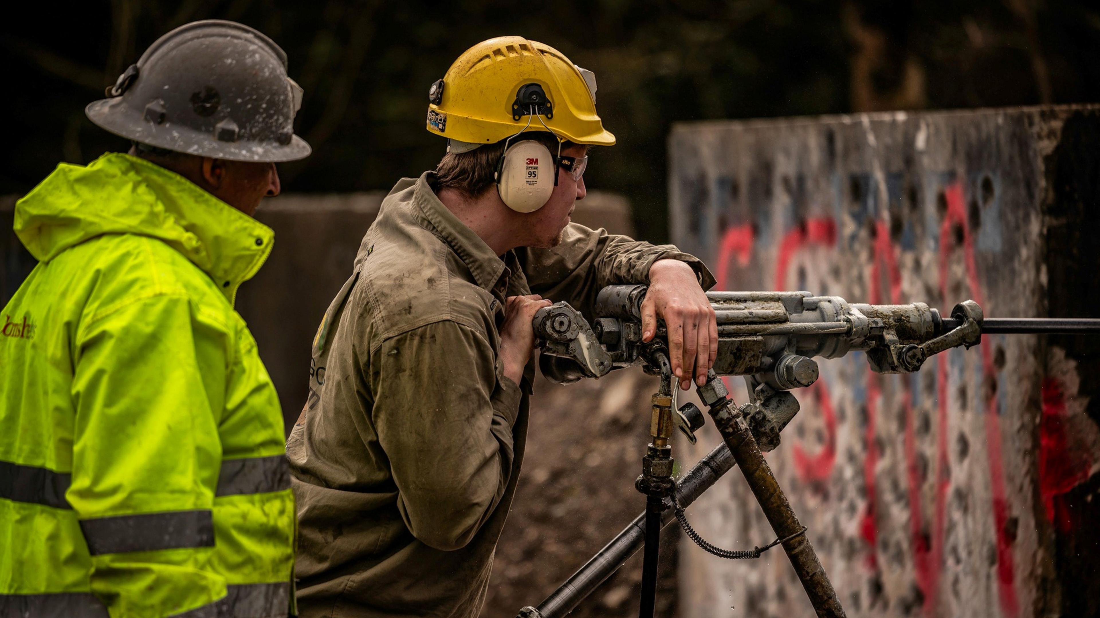 Two people with a mining drill. One person is using the drill, drilling into a wall while the other is observing. The observer is wearing a high viz jacket with a hardhat while the person using the drill is in brown overalls. 