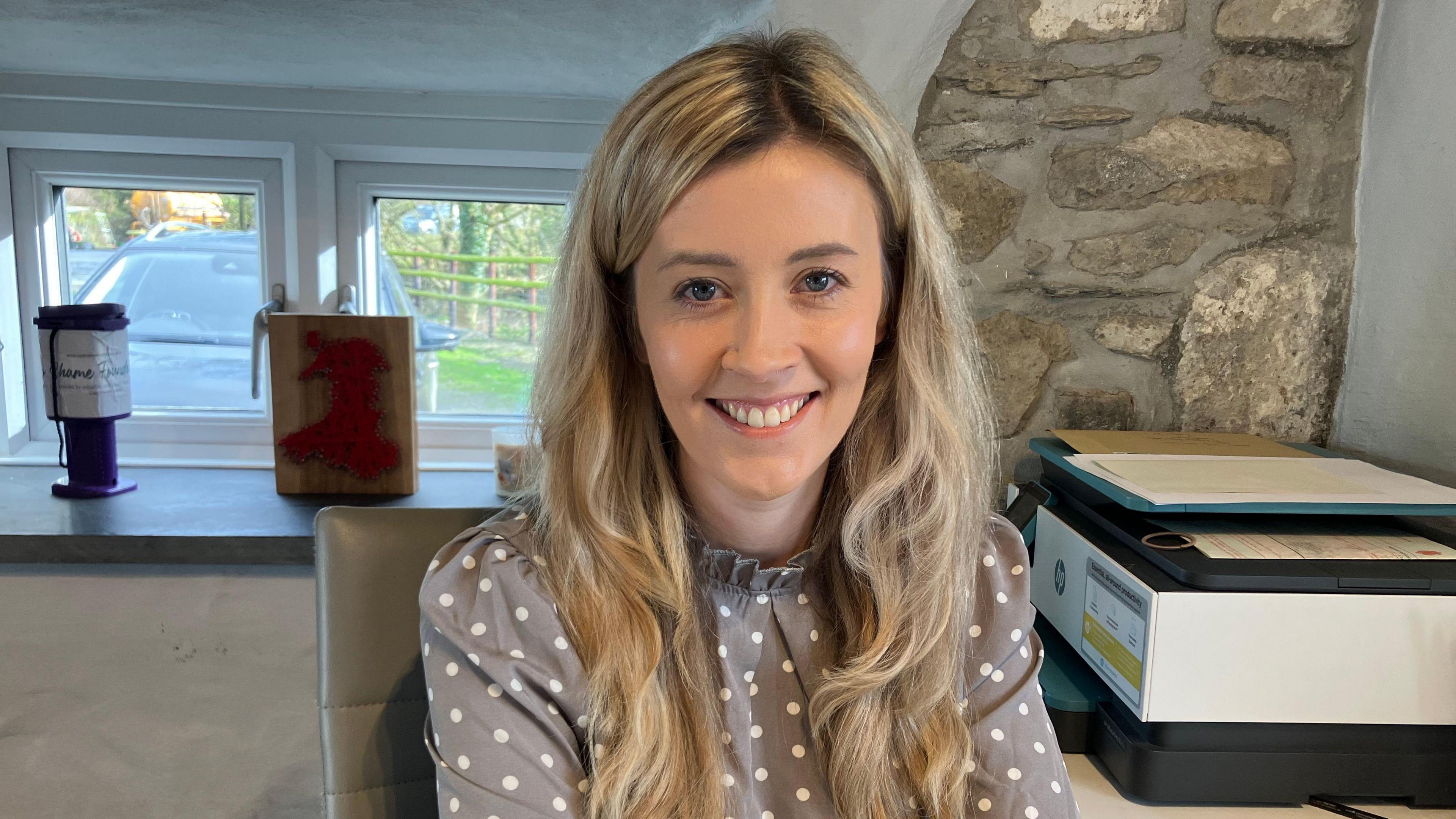 Glesni Evans yn gwisgo crys patrymog llwyd, gwallt hir melyn, yn gwenu.