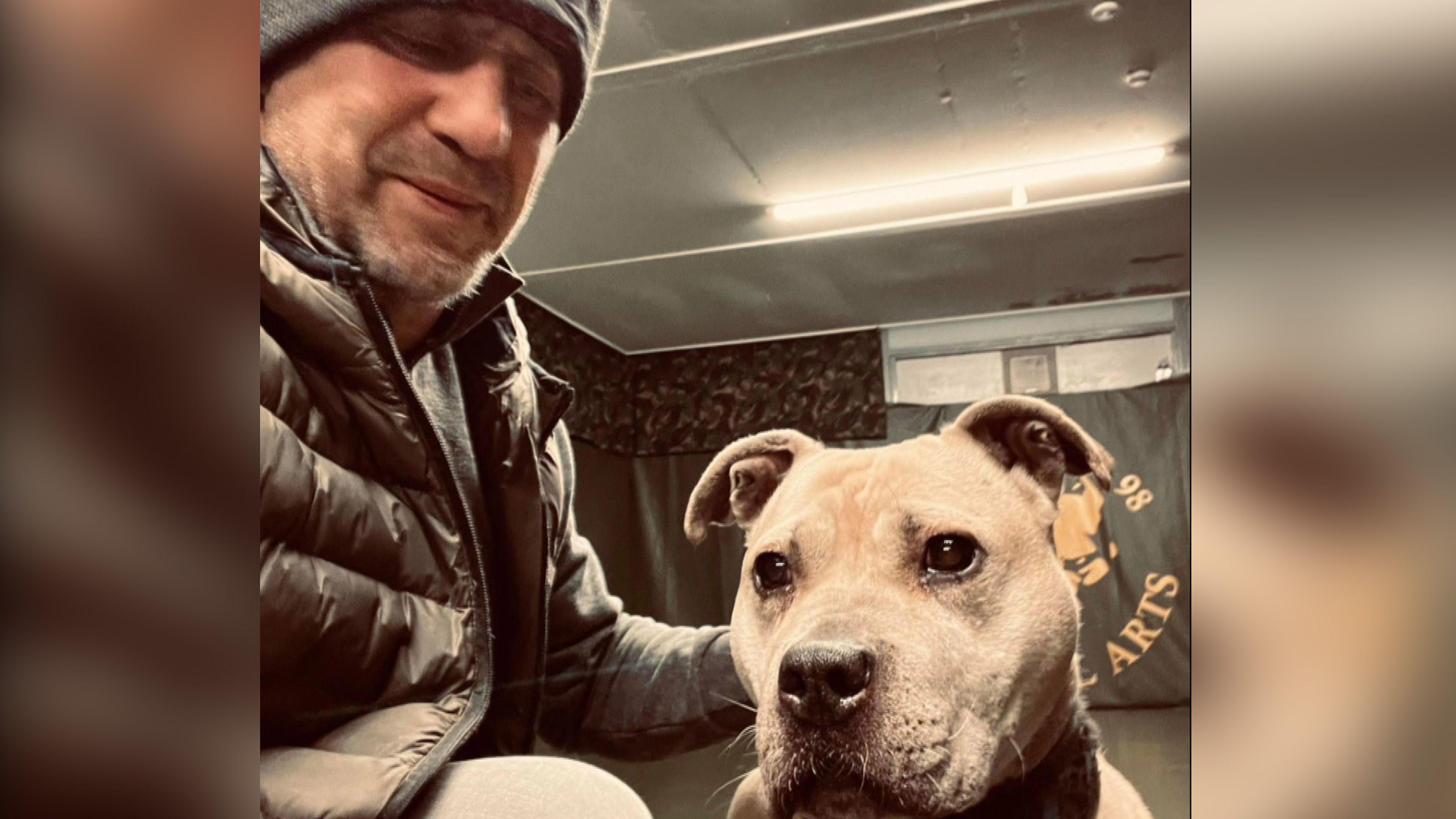 A man in a black coat and wooly hat is crouched down in a gym training room with a pitbull type dog. He has a dark stubble which is turning grey. The dog is a tanned colour and is staring intently to the left of the camera.  Behind them is a large gym banner. 