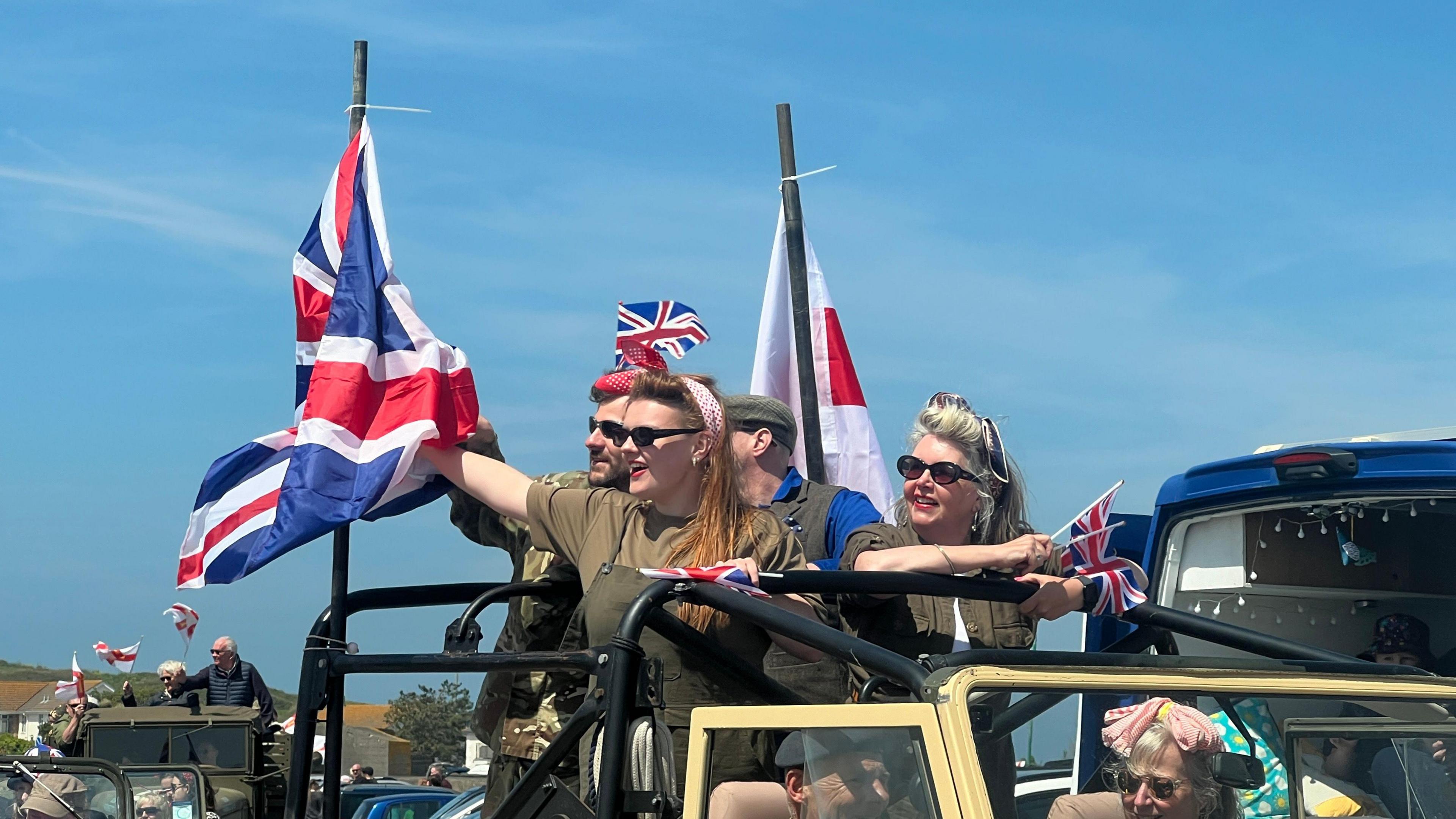 Men and women riding in a car during the Liberation Day 2024 cavalcade. They are wearing sunglasses, khaki outfits and are waving Union Jack flags.