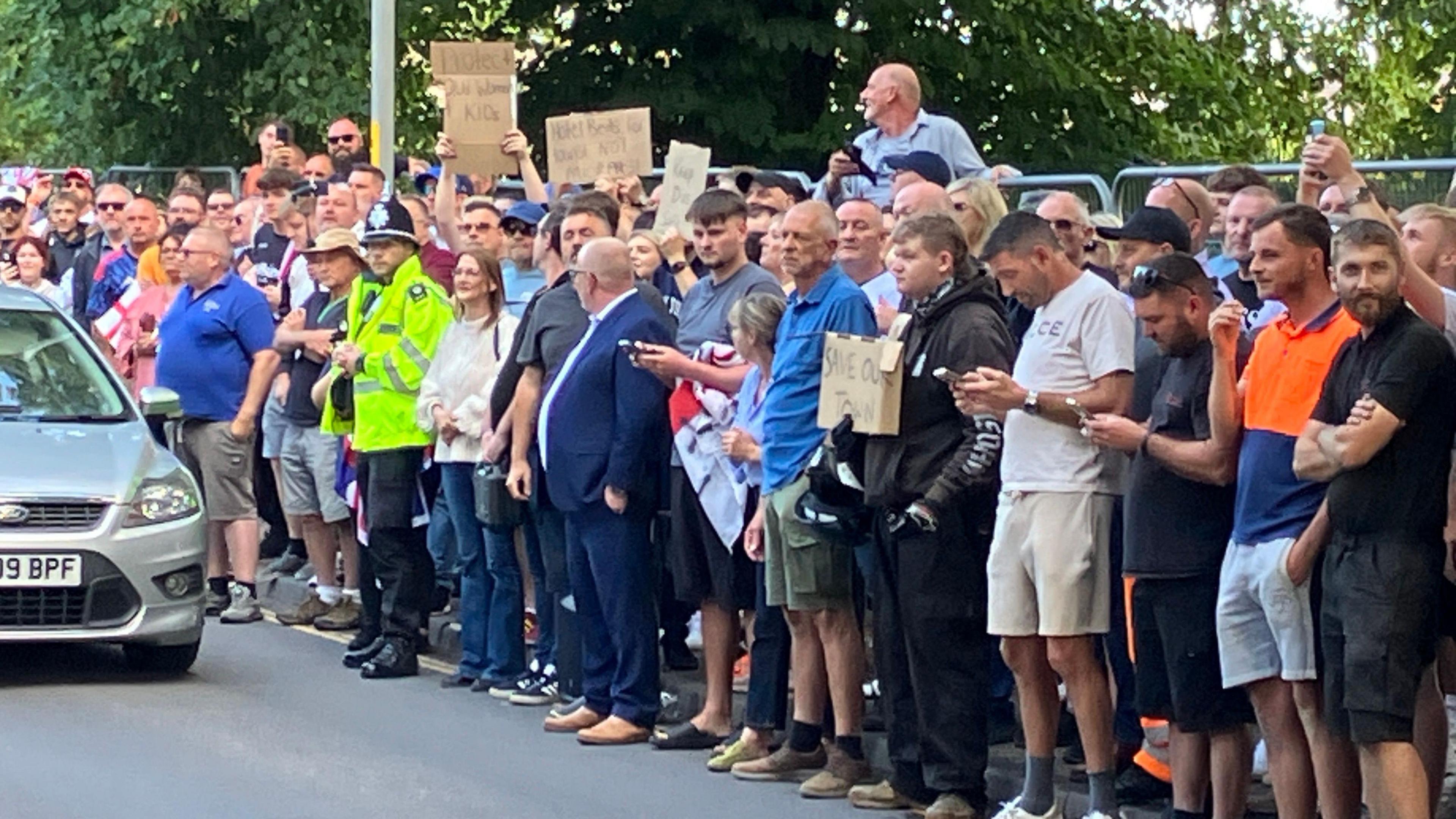 A large group of protesters have overfilled the pavement and spilt onto the road. Some are holding signs. A police officer is stood with them.