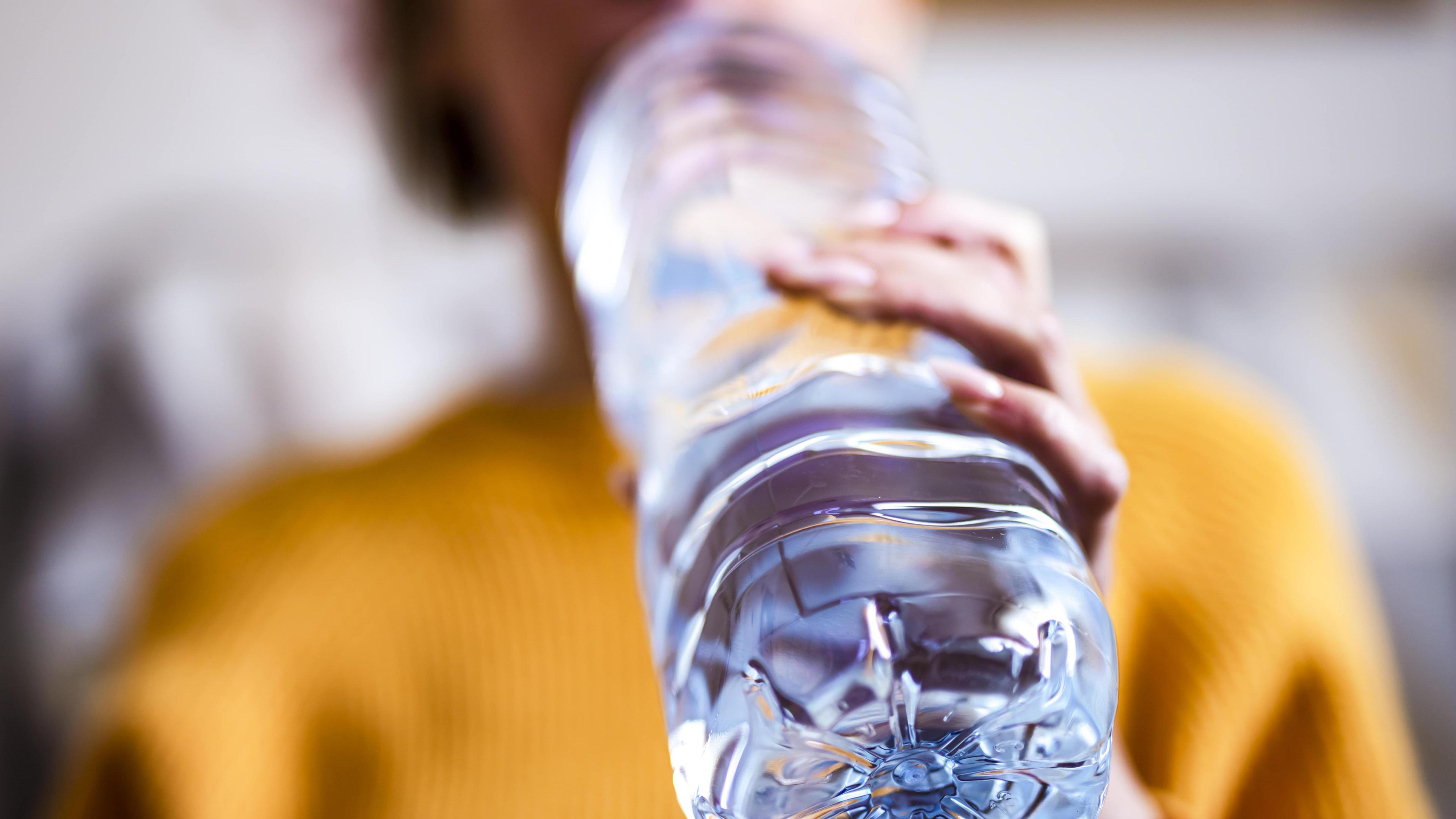 A woman wearing a mustard coloured top takes a drink from a large unlabelled bottle of water. The background is blurred maintaining focus on the bottle. 