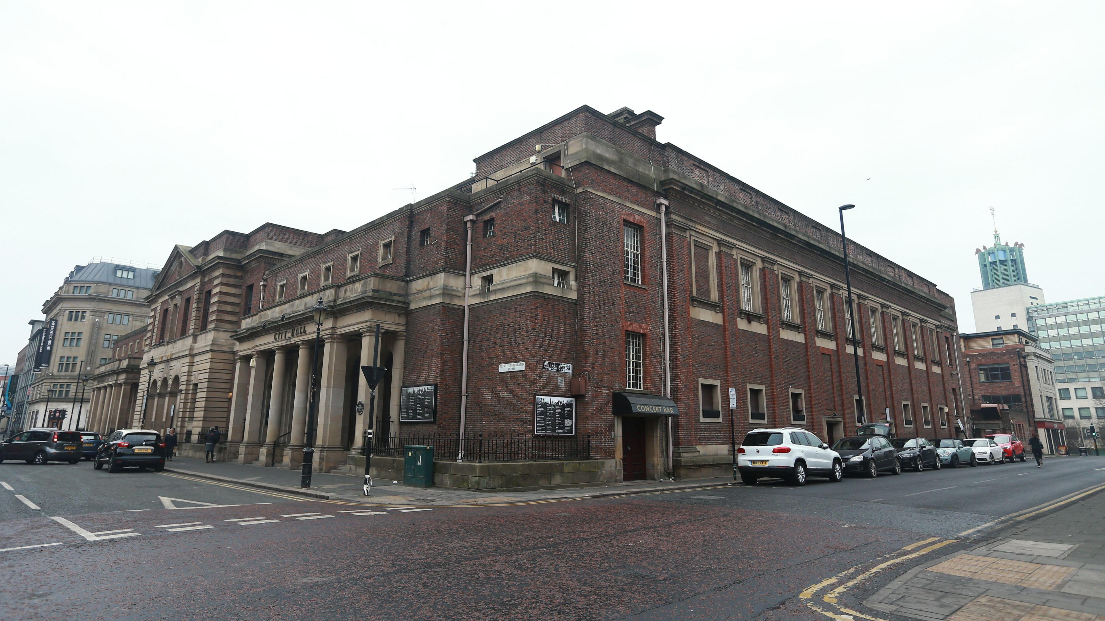 A picture of Newcastle City Hall. The photograph is taken from the corner of the street so two sides of the red-brick building are in view. The entrance stands to the left and has large sandstone pillars in front of the door. The City baths are further to the left.