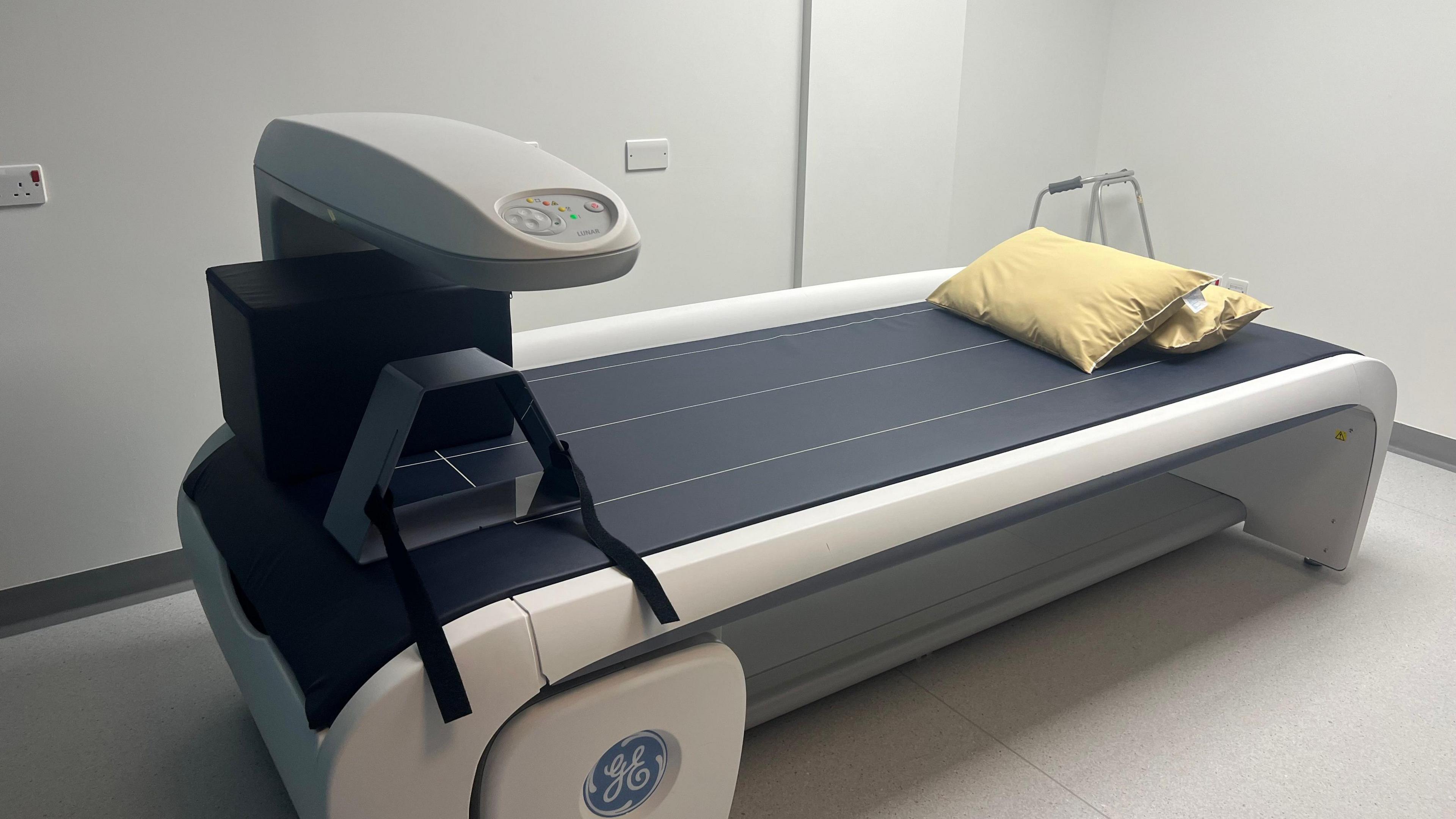 A bone density scanner in a room with white walls. The scanner is like a long hospital bed without sides. There are pillows on one side and a plastic section that comes up over the bed with buttons.