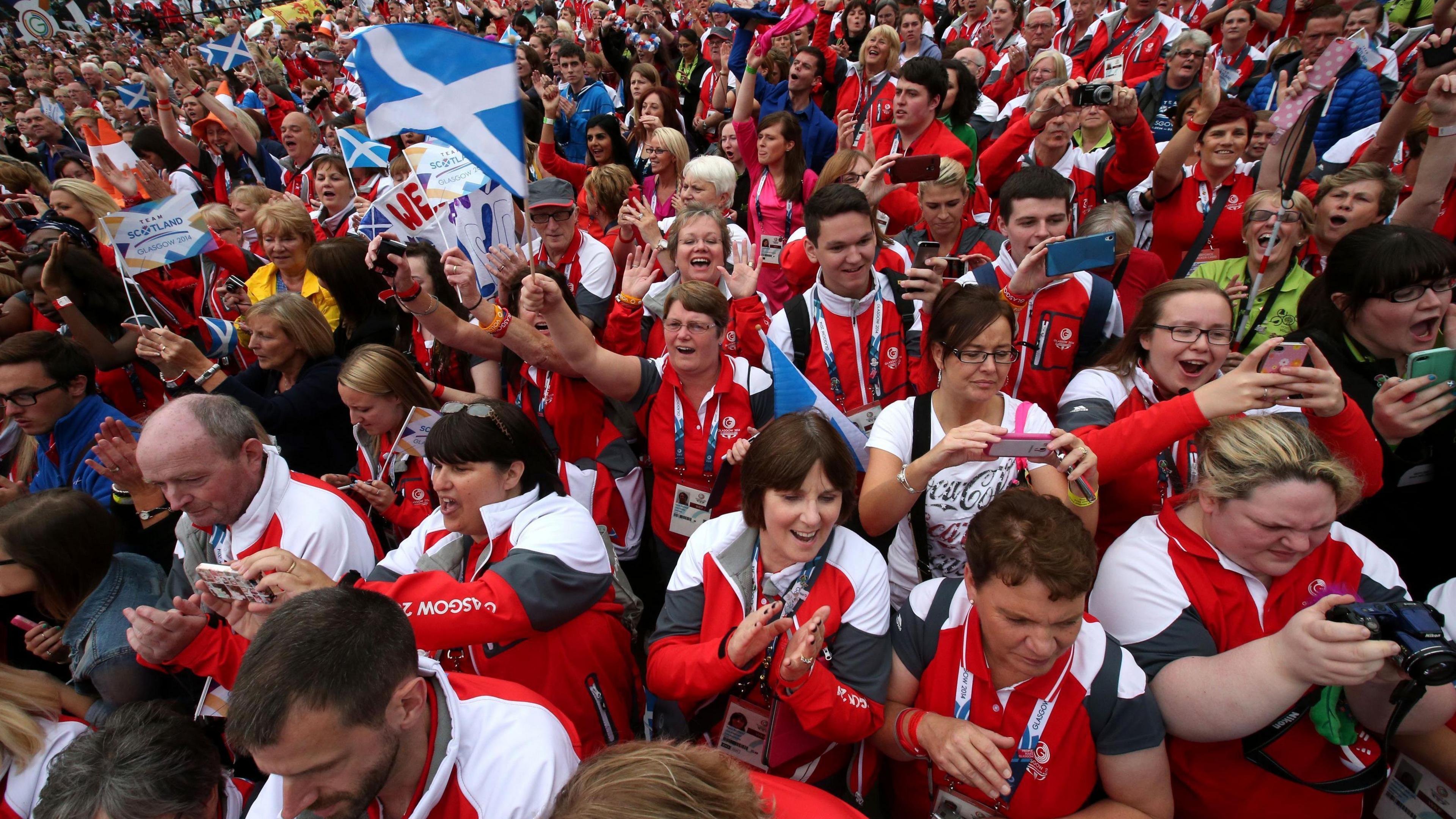 A parade of volunteers from the 2014 Commonwealth Games. They are all wearing red and white uniforms, and many of the dozens pictured are cheering