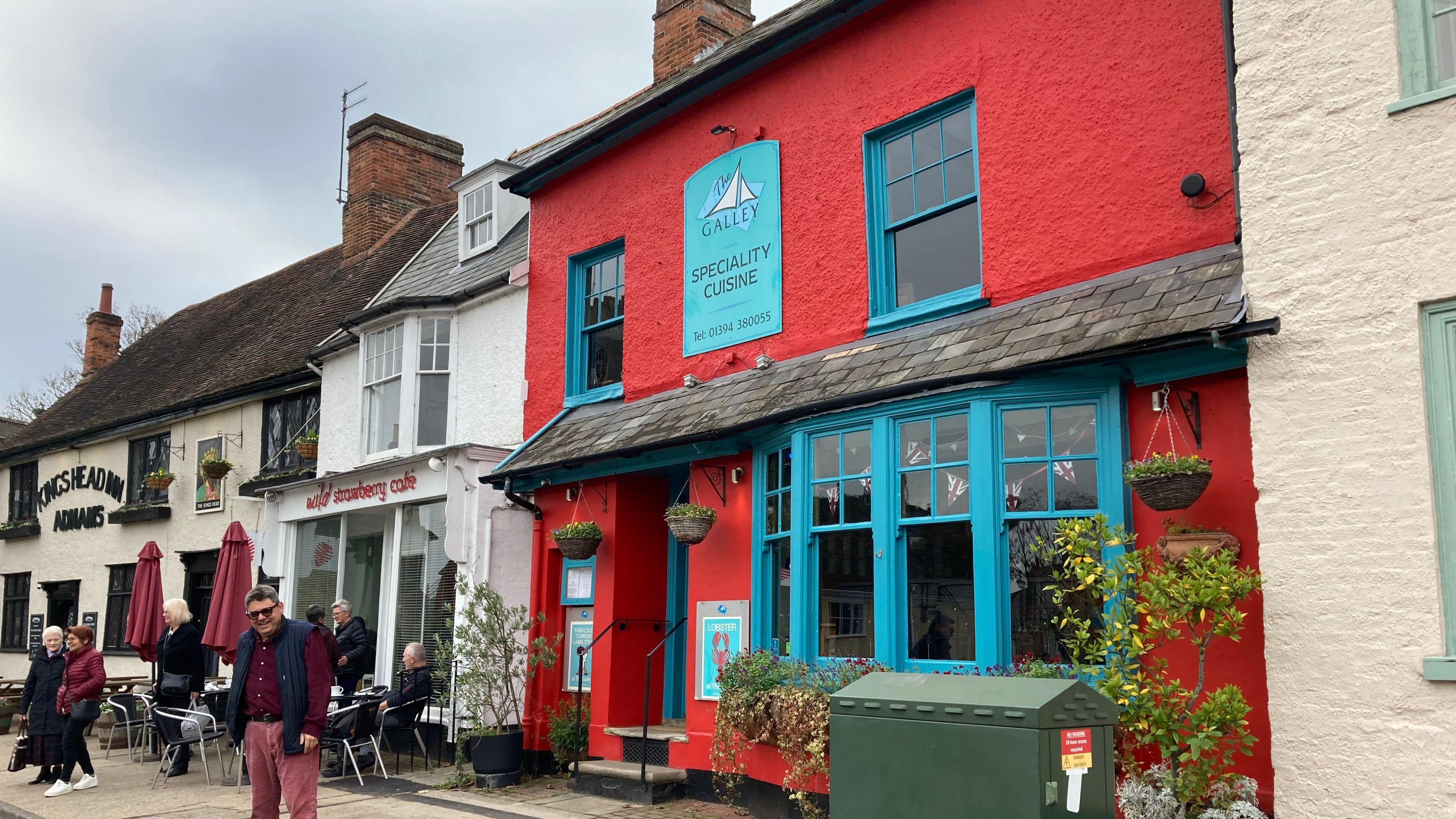 Mr Vata stands outside his restaurant. He is smiling to the camera. People can be seen leaving a cafe next door. The Galley is a bright red building with teal coloured window frames and a door. Plant pots hang from the building and windows.