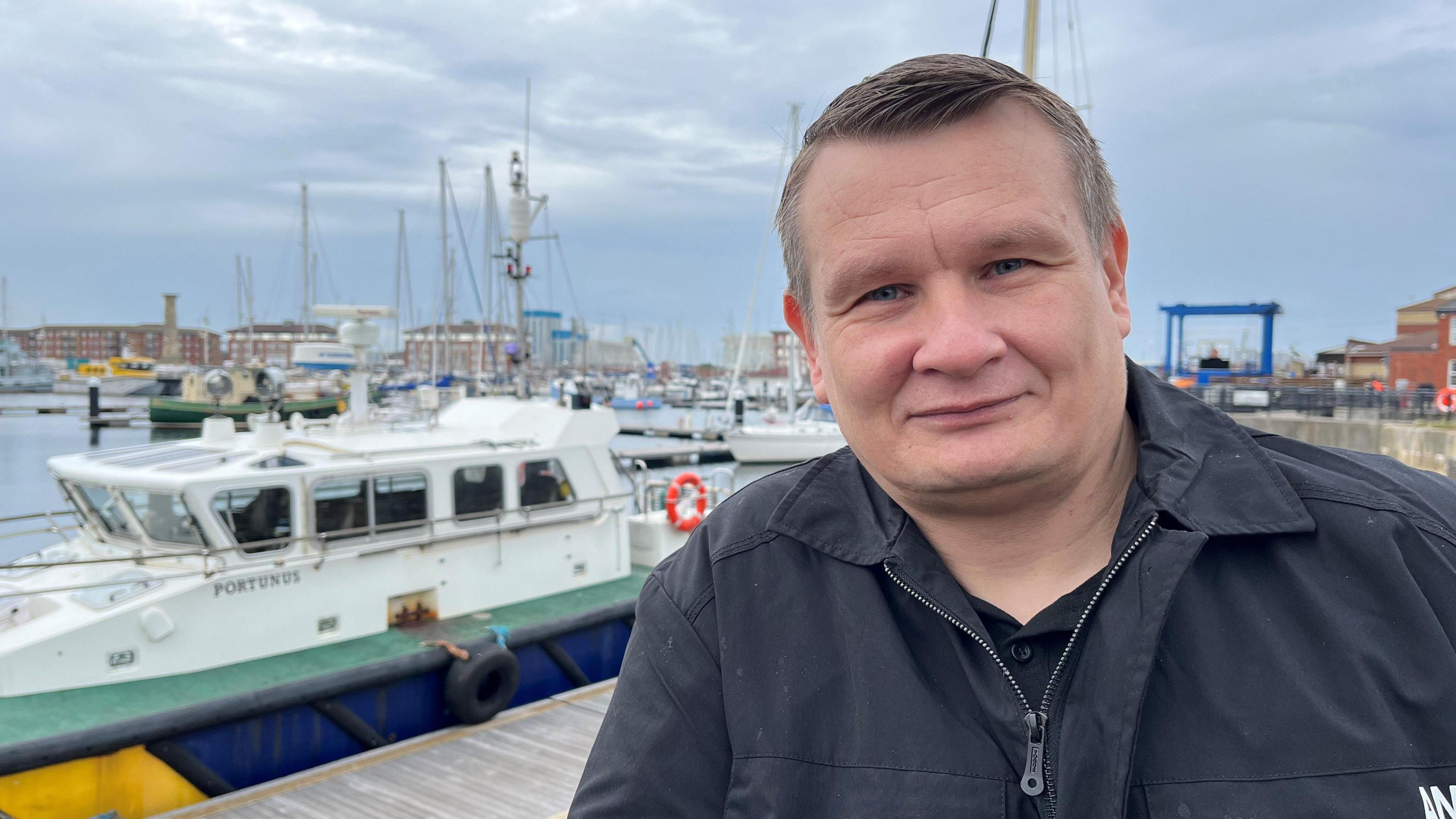 A man with short hair and a black jacket standing at Hartlepool Marina with water and a moored boat behind him