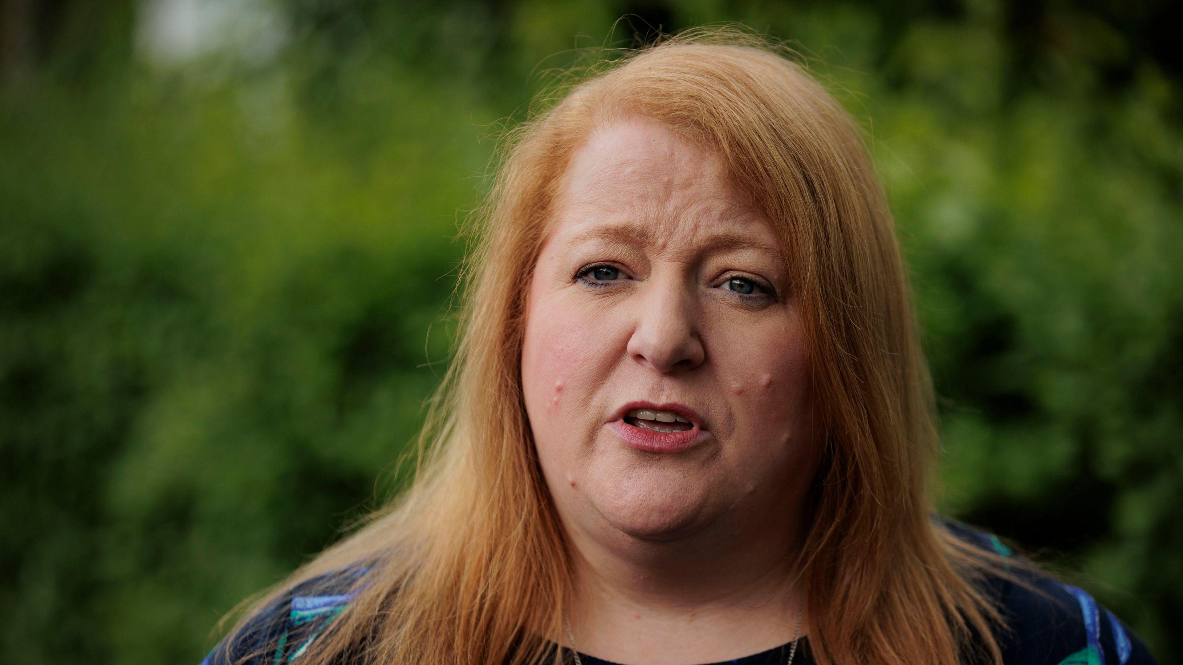 An up close image of Naomi Long. She has strawberry blonde hair and has a serious look on her face. She is wearing a navy top. There are blurred green bushes in the background. 