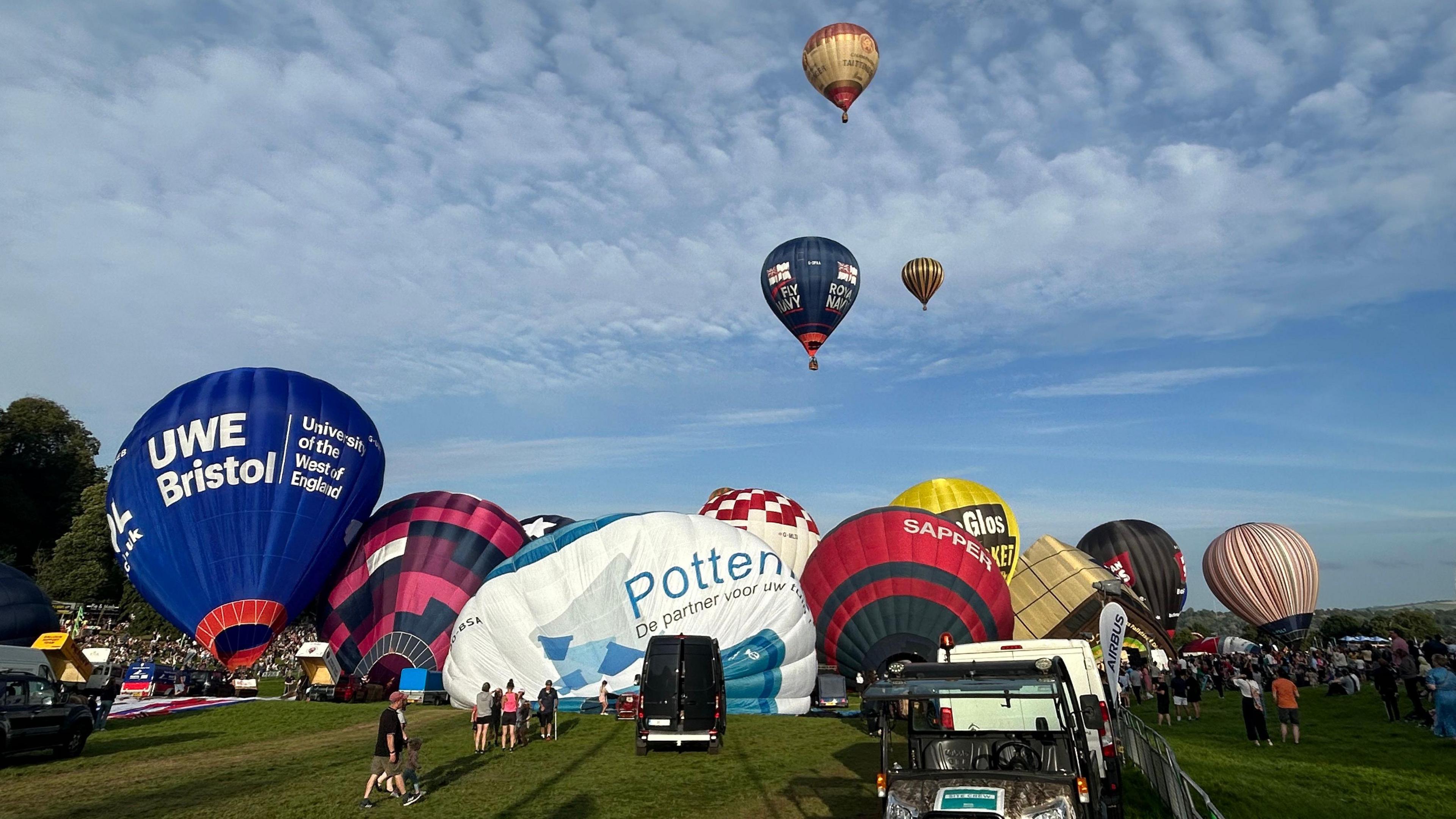 Hot air balloons inflating and taking off at Ashton Court. The balloons are many different colours and patters, some with branding on them. 