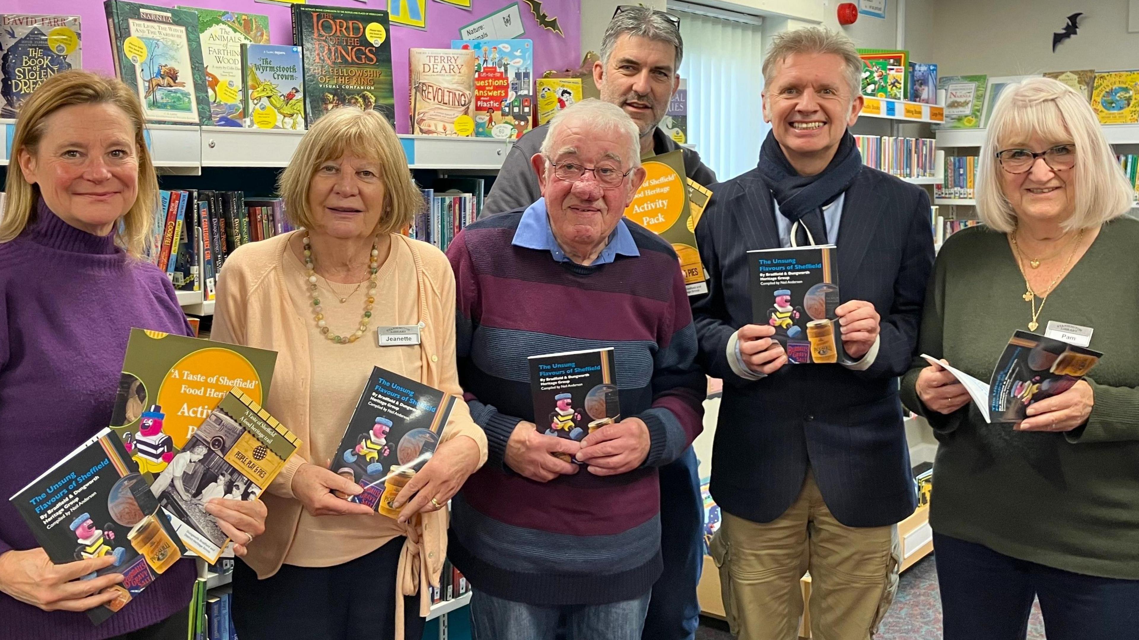 Six people in a line. They hold up copies of the same book and are all smiling at the camera. They are in a library with a shelf full of colourful books is behind them.