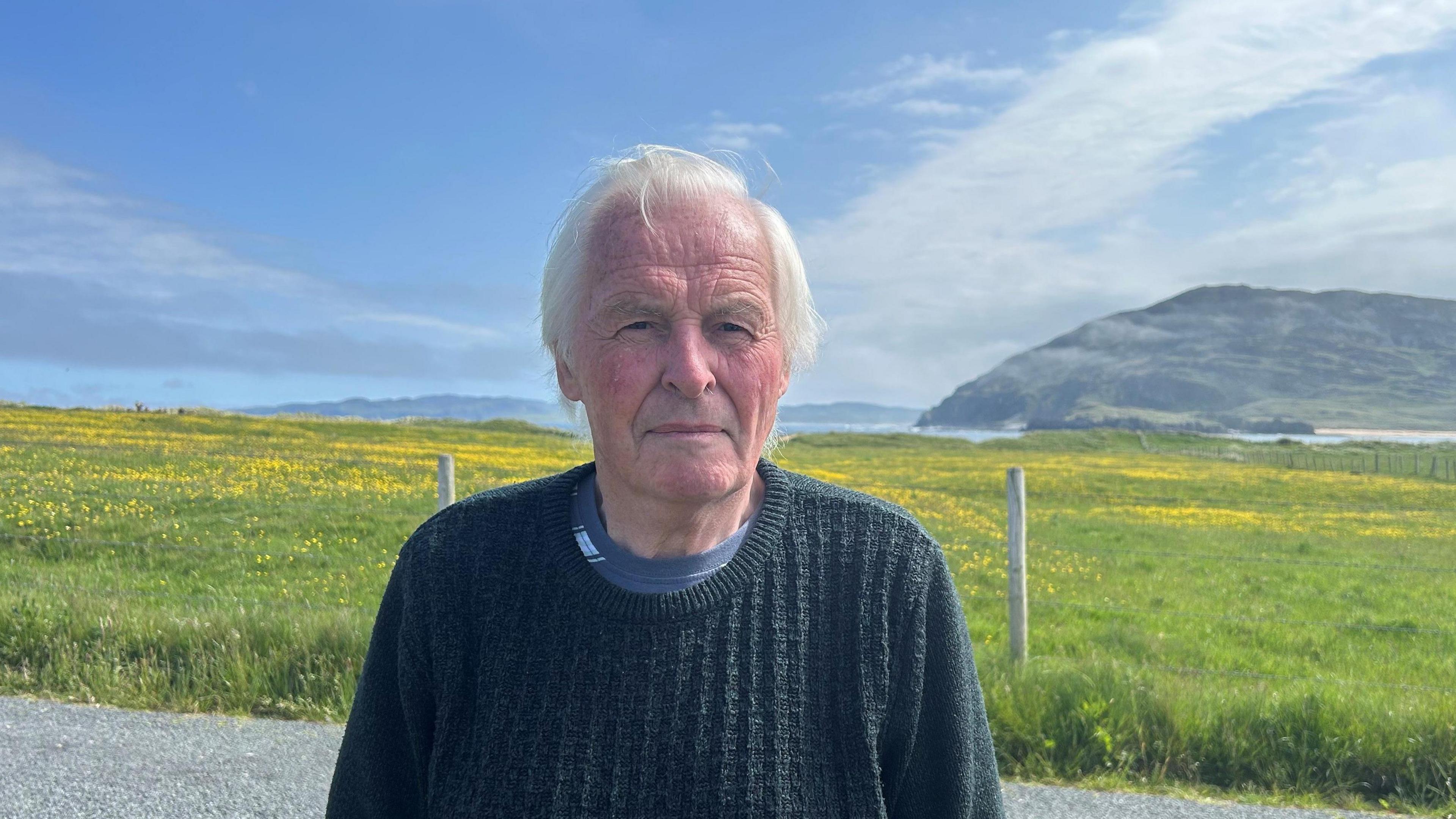 A man in a thick knitted jumper is standing in front of a field, filled with tall grass and small yellow flower heads. Behind him is a coastline and a mountain, with sprawling blue skies. The man has flowing grey hair and is looking directly at the camera. 