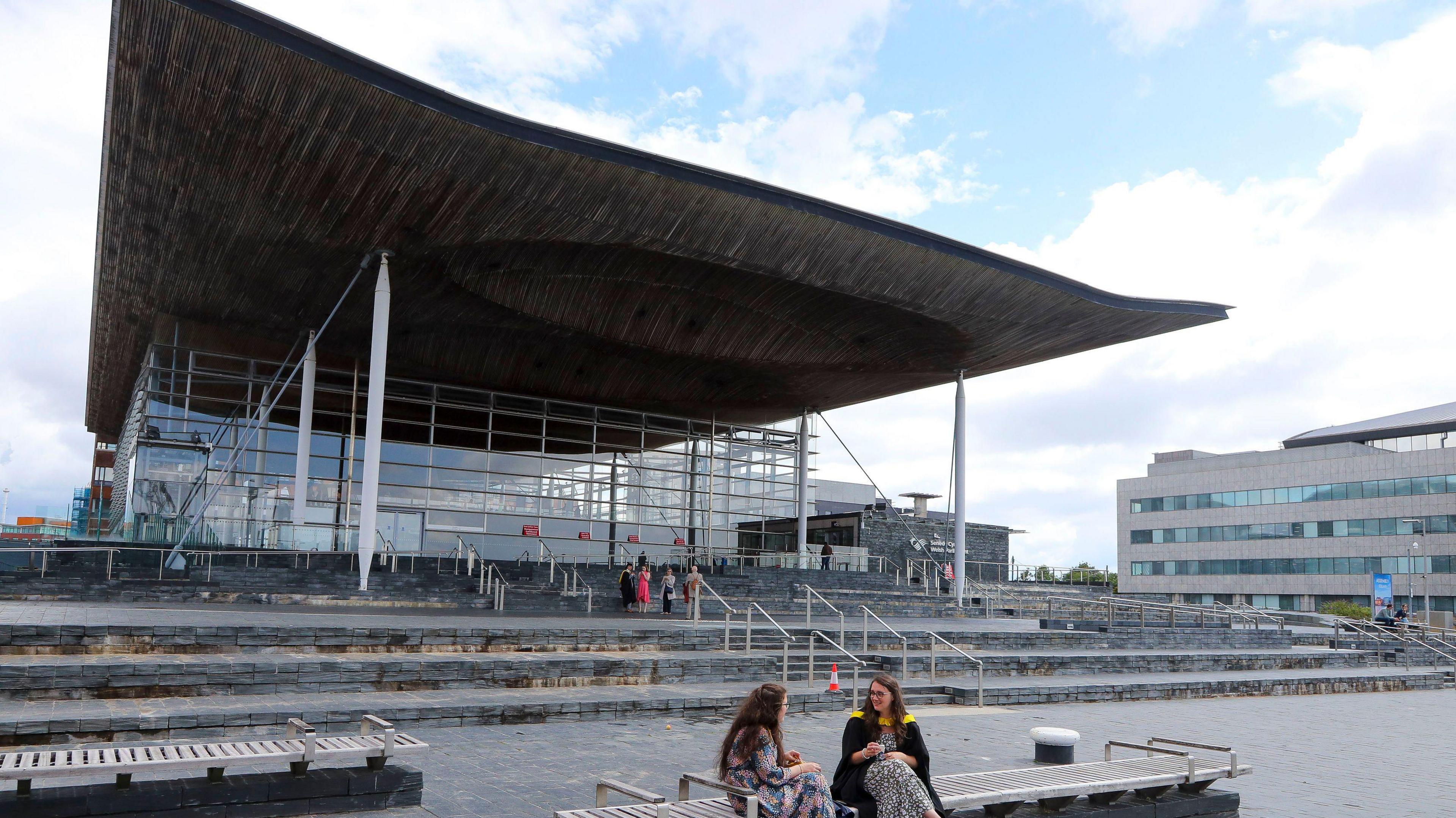 A picture of the Senedd from the front, facing into the rest of Cardiff Bay. The steps, which are clad in slate, can be seen, with two women sat on a bench in view.