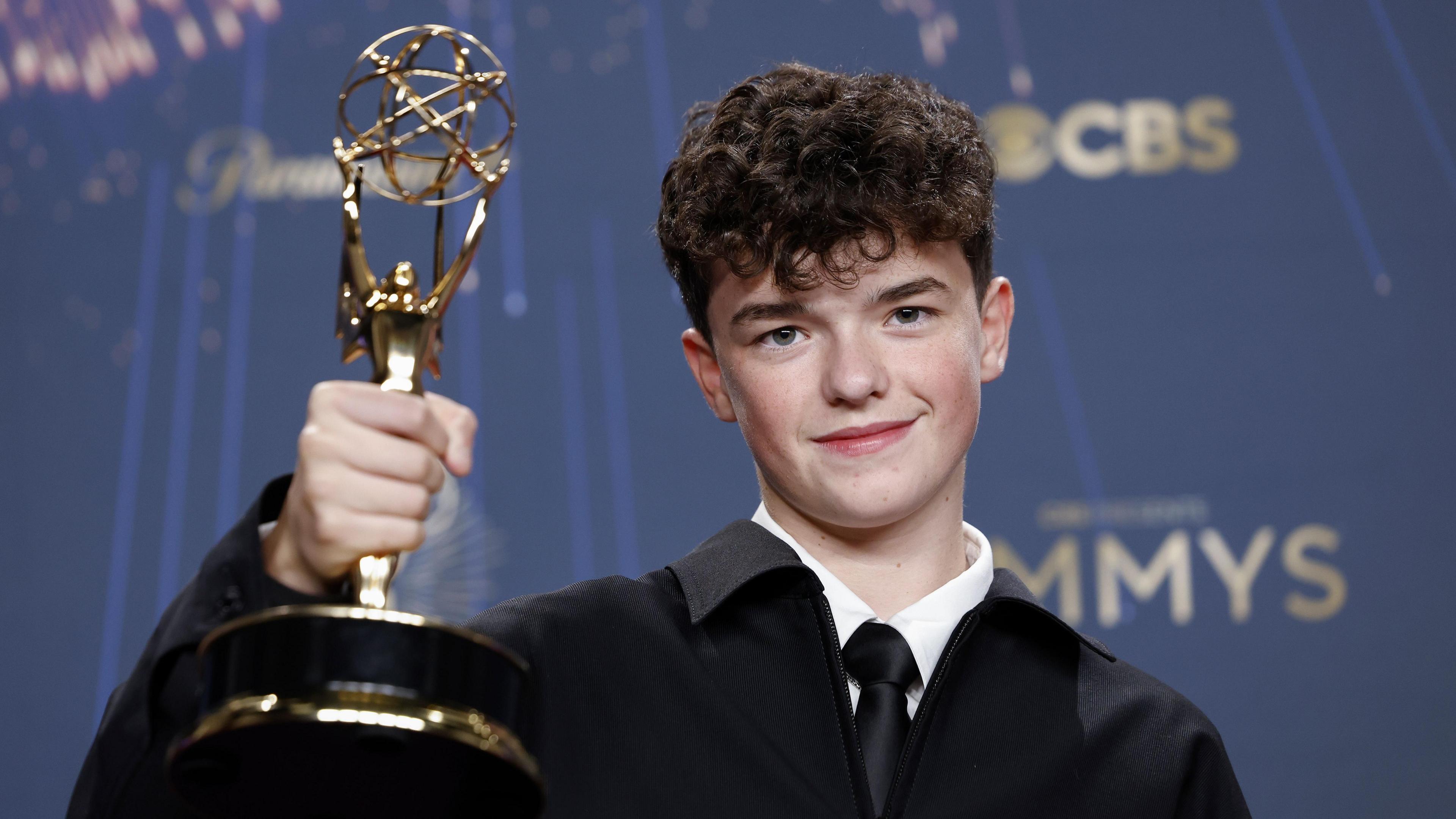 Owen Cooper smiles and holds up his Emmy award in front of a dark blue backdrop. He wears a black suit with a white shirt and black tie.