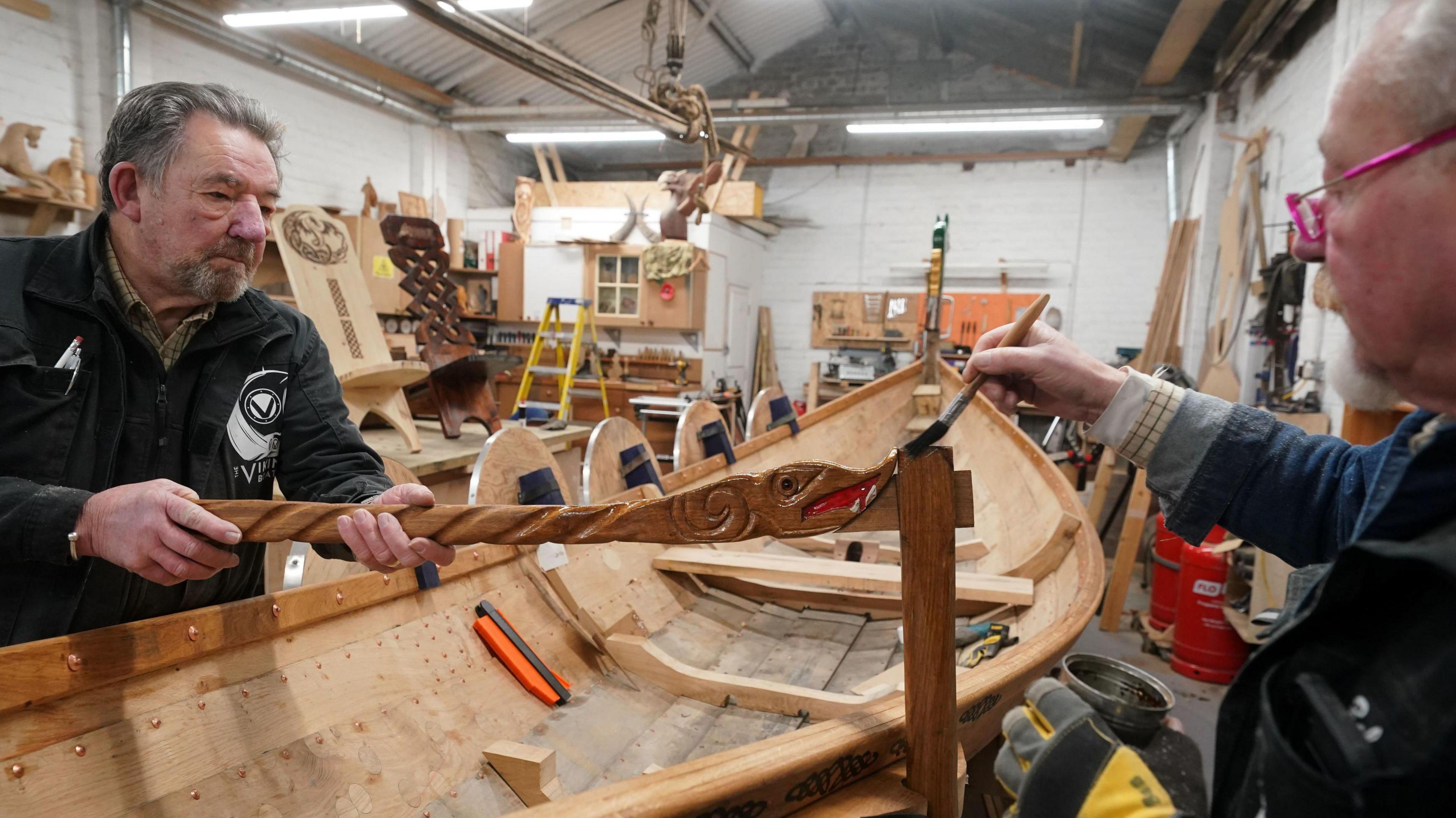 Two men are painting a piece of wood carved into the shape of a dragon on either side of a Viking boat. They are both older men in their 60s or 70s.