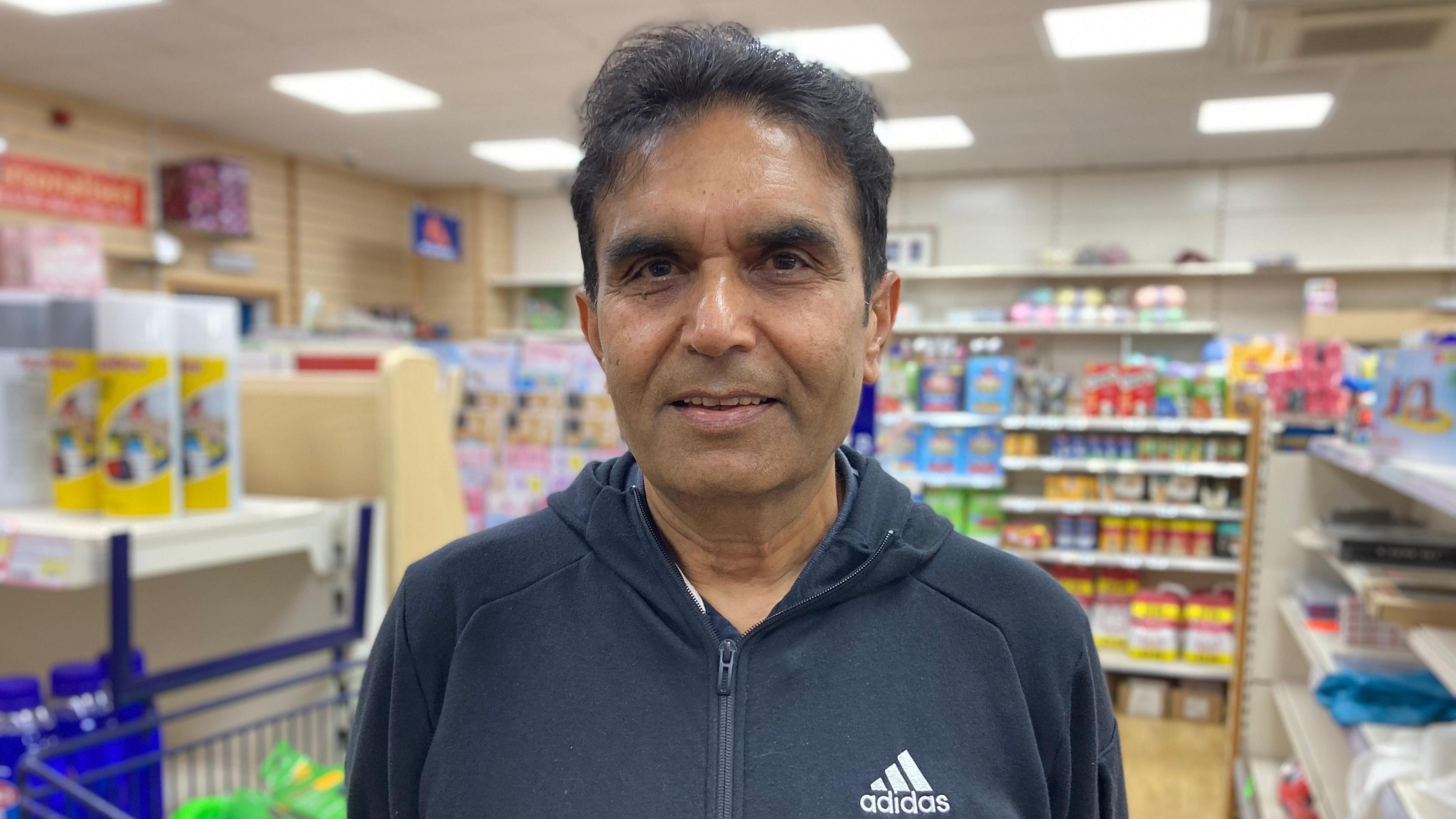 Danny Hirani, wearing an Adidas zip up dark hoody, standing in a convenience store