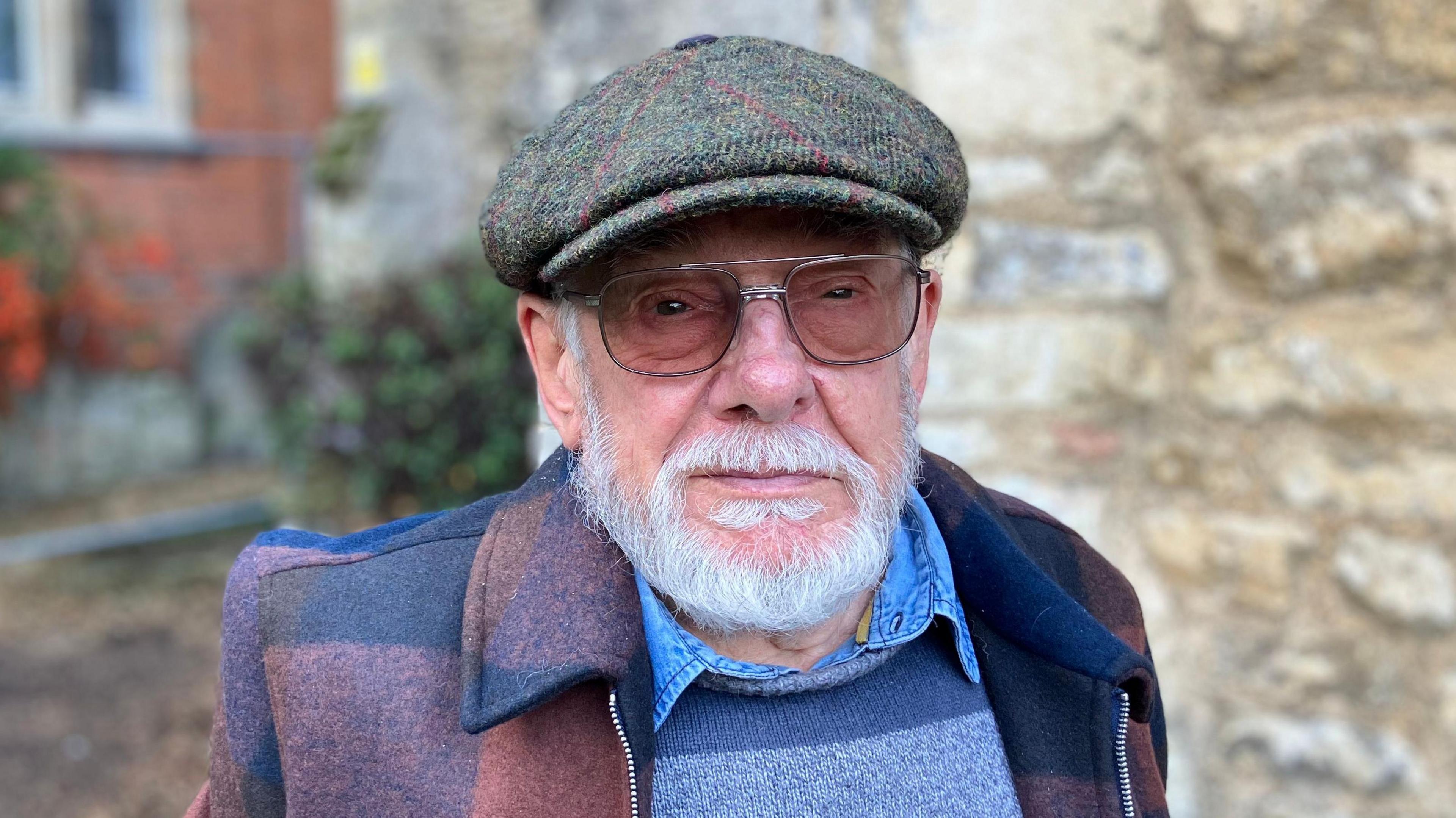 Colin Hardy is in the middle of the image looking towards the camera. He has a short grey beard and is wearing a glasses, a green tweed hat, blue shirt and jumper and a chequered coat. 