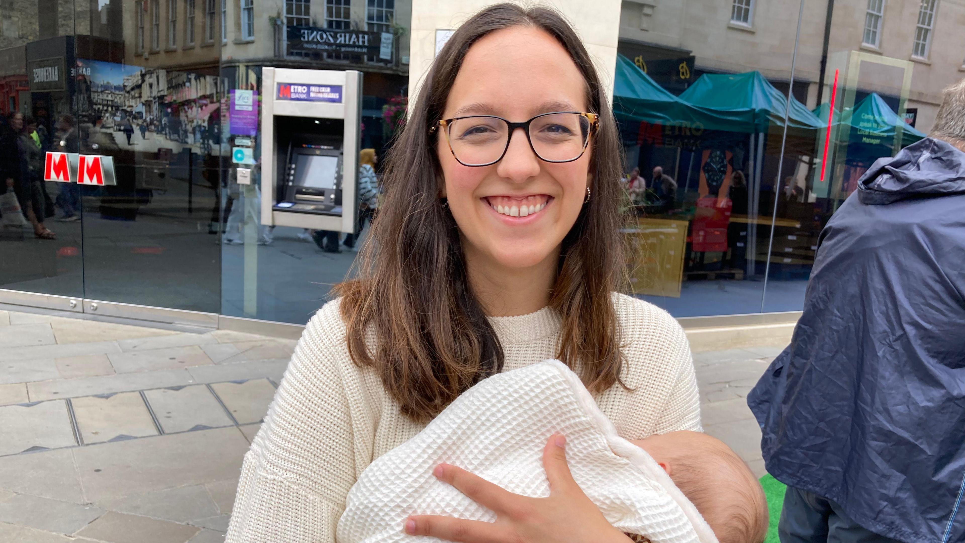 Carmen Murillo standing in the street holding her three month old daughter, who is wrapped in a white blanket. Carmen has mid-length brown hair and is wearing thin framed glasses and smiling, wearing a knitted cream jumper