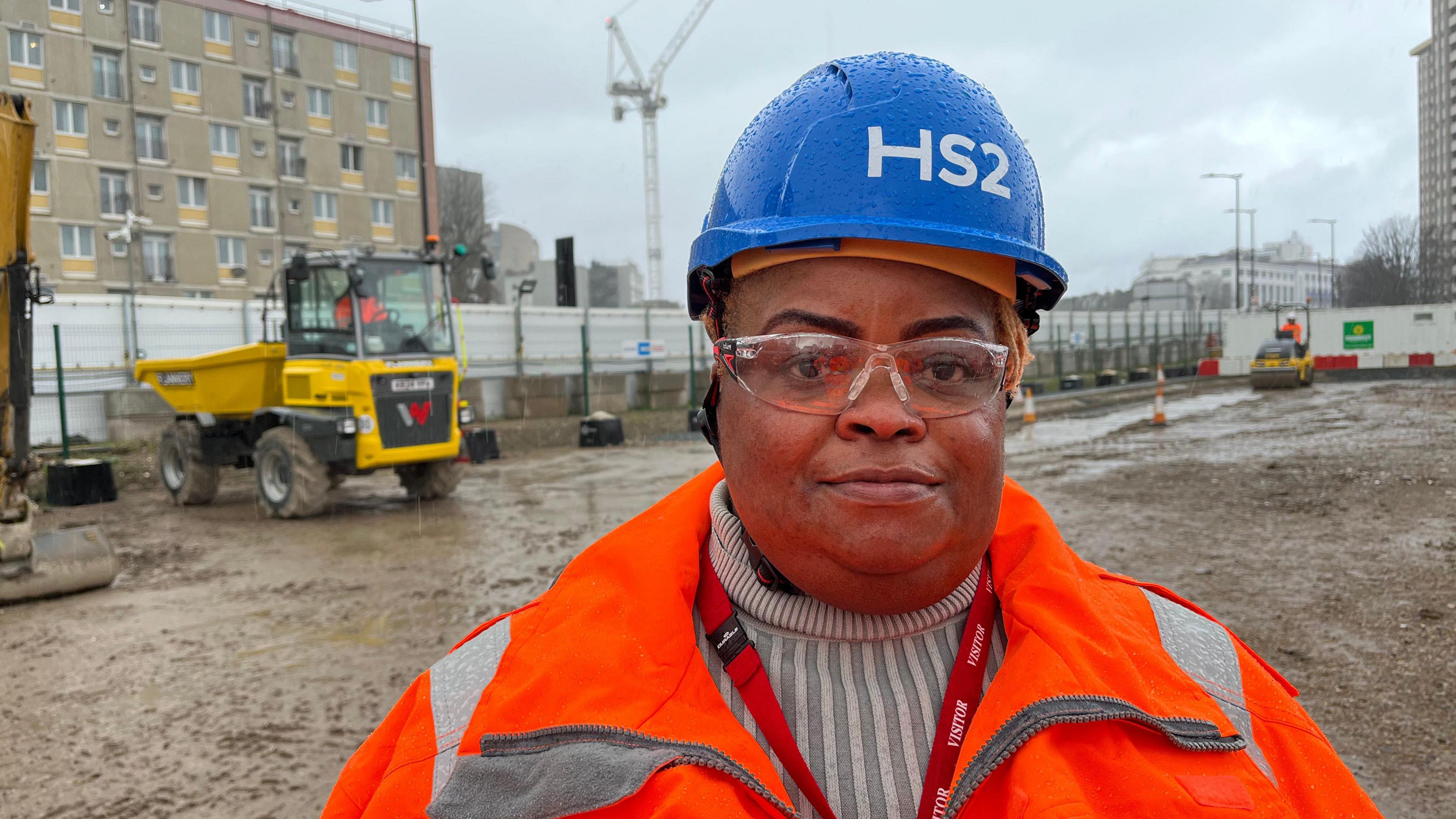 Beatrice Adegbola is looking into the camera. She is wearing a blue hard hat and orange safety jacket. In the background is a muddy area with a digger and a crane