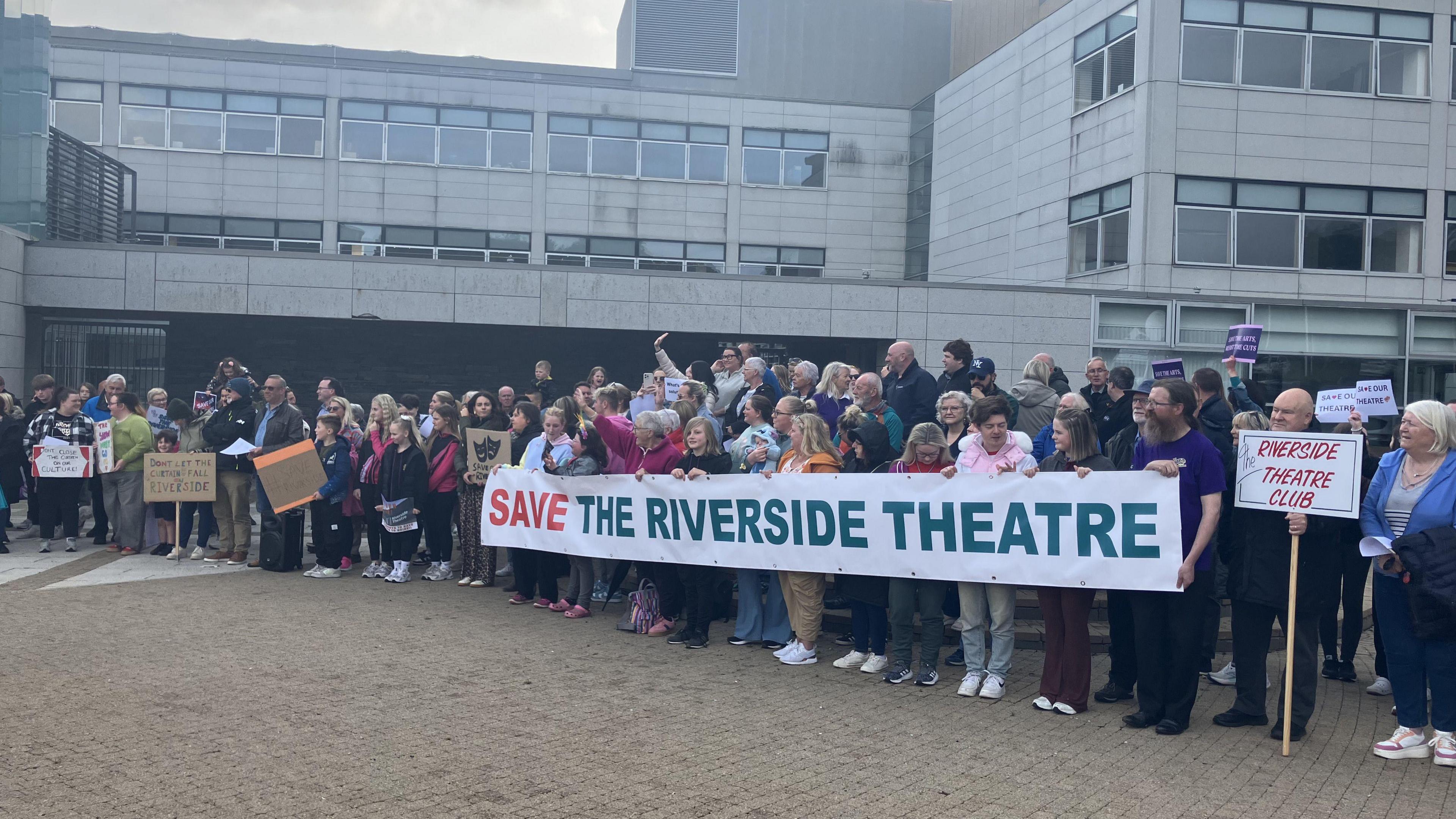 A crowd of approximately 150 people are outside council offices protesting. They have a banner and placards reading "save the riverside theatre". The crowd is made up of young and older people