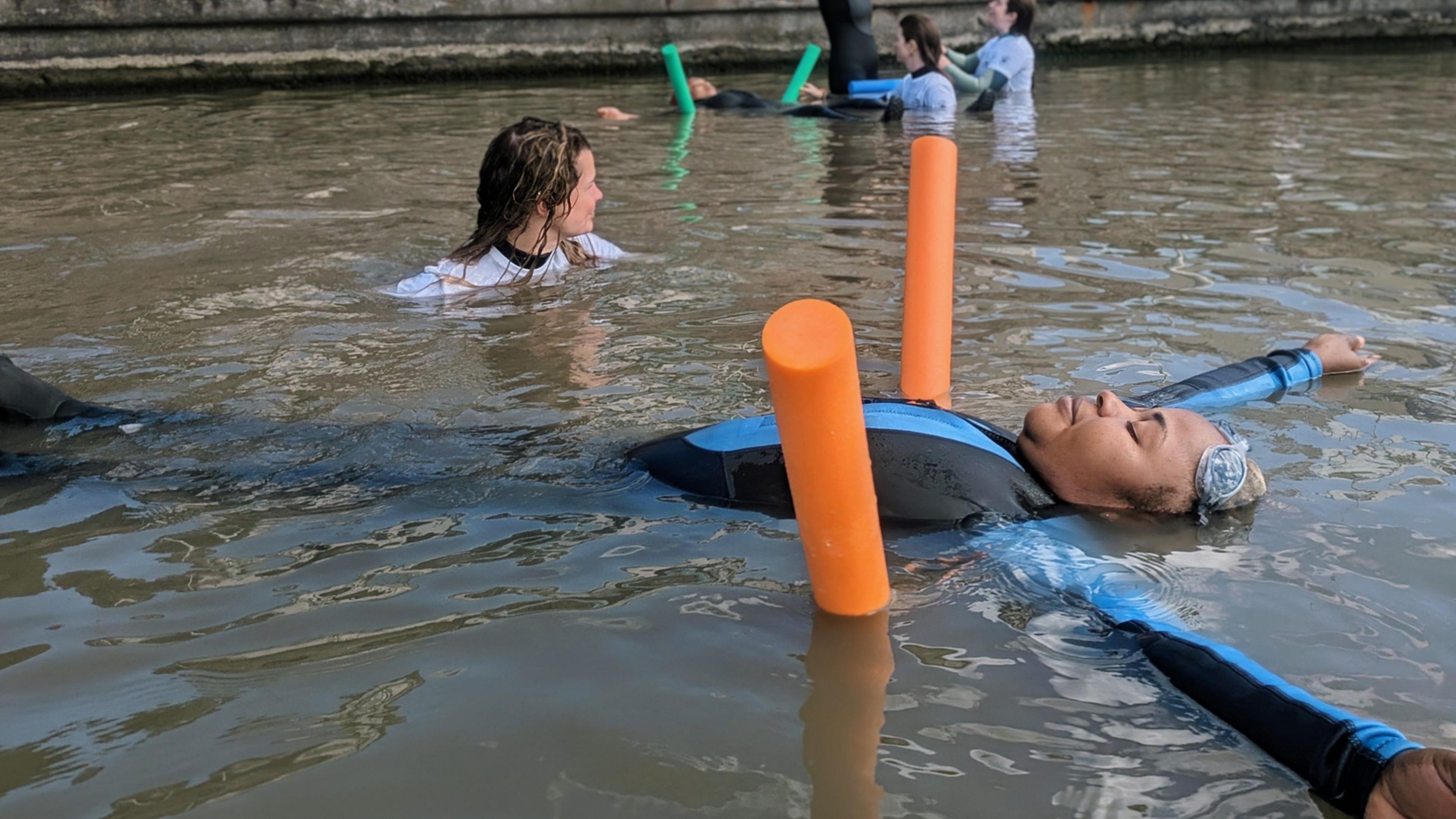 A woman wearing a blue and black wetsuit and goggles. She is floating on her back in Weston-super-Marine Lake with her eyes closed. An orange pool noodle is underneath her back, helping her stay afloat