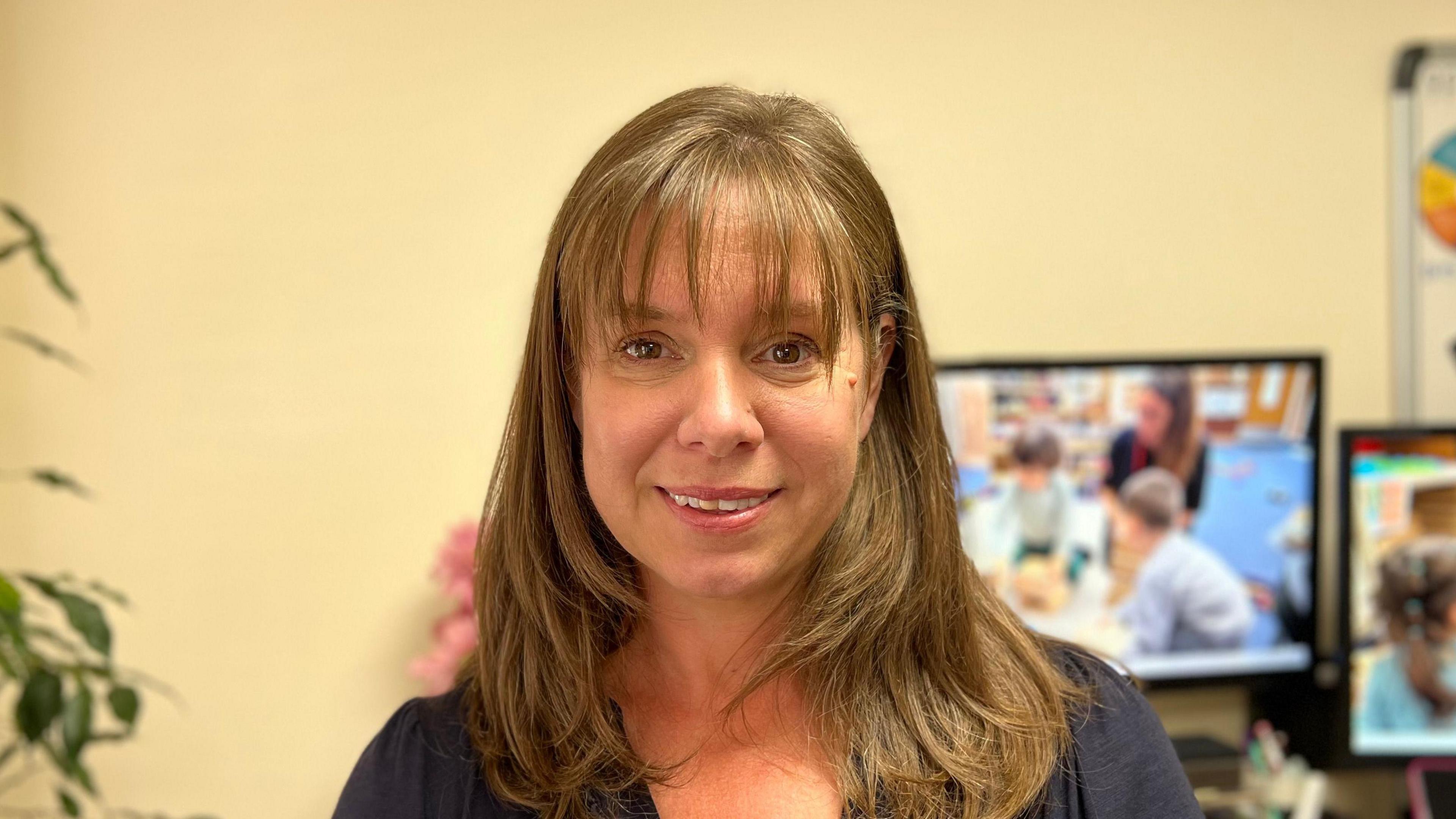 Fiona Vacher is standing in her office. She is looking into the camera. Behind her are cream walls and two computer screens, the desktop backgrounds are children at a playgroup.