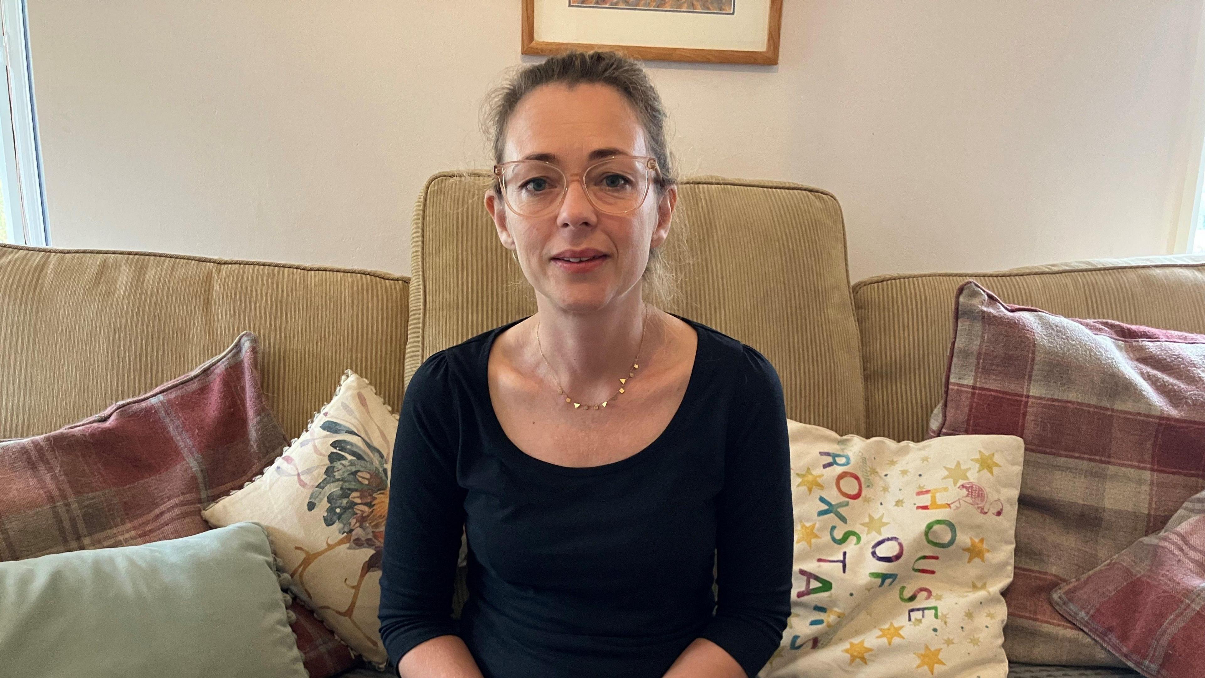 A woman in a black top wearing glasses. She is sat on a sofa with lots of colourful cushions on it.
