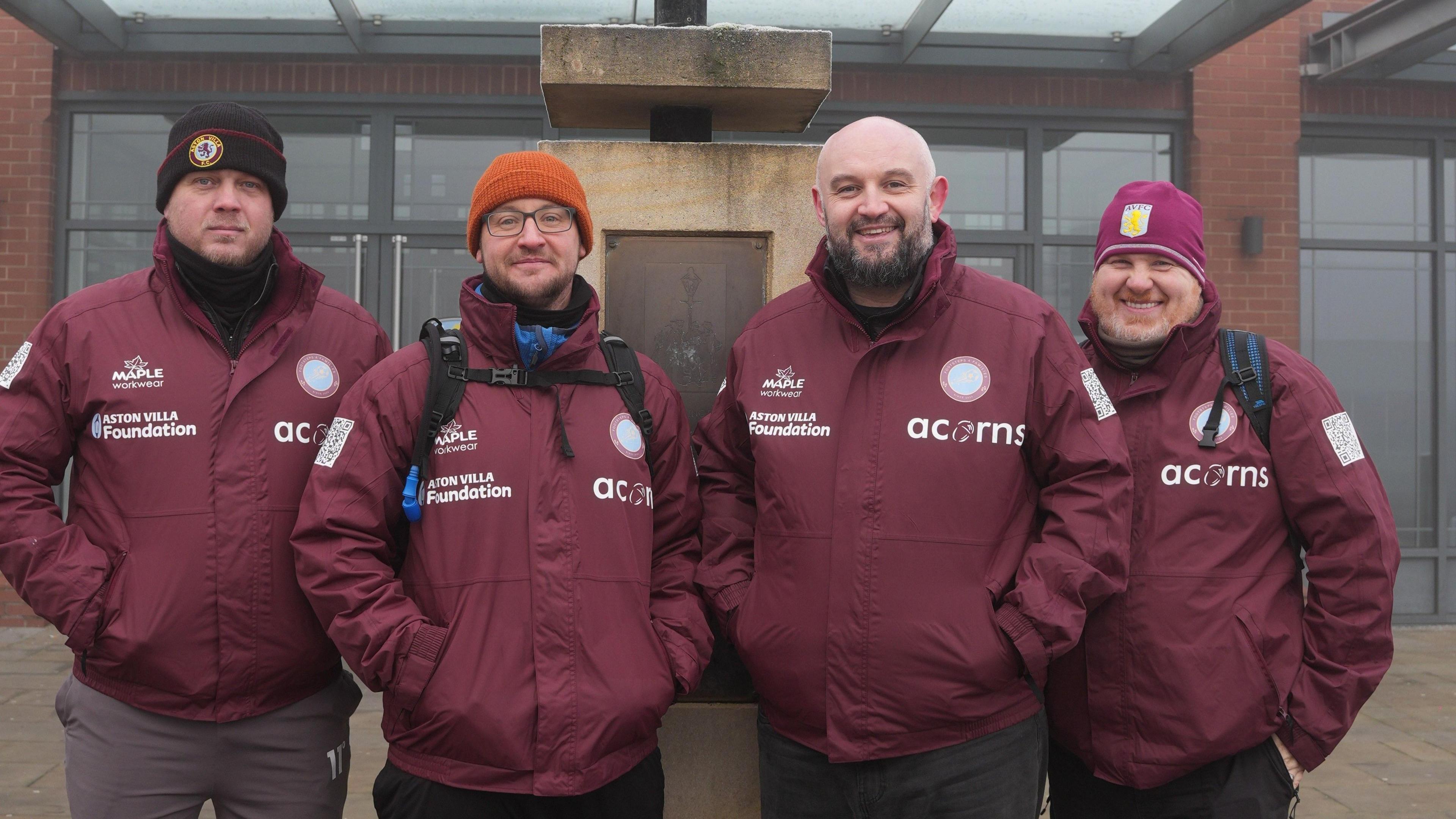Four men standing in a line. They are all wearing maroon waterproof jackets with the acorns children's hospice logo on the front, as well as the Aston villa foundation logo. Three of the men have beanie hats on, and one has a bald head.