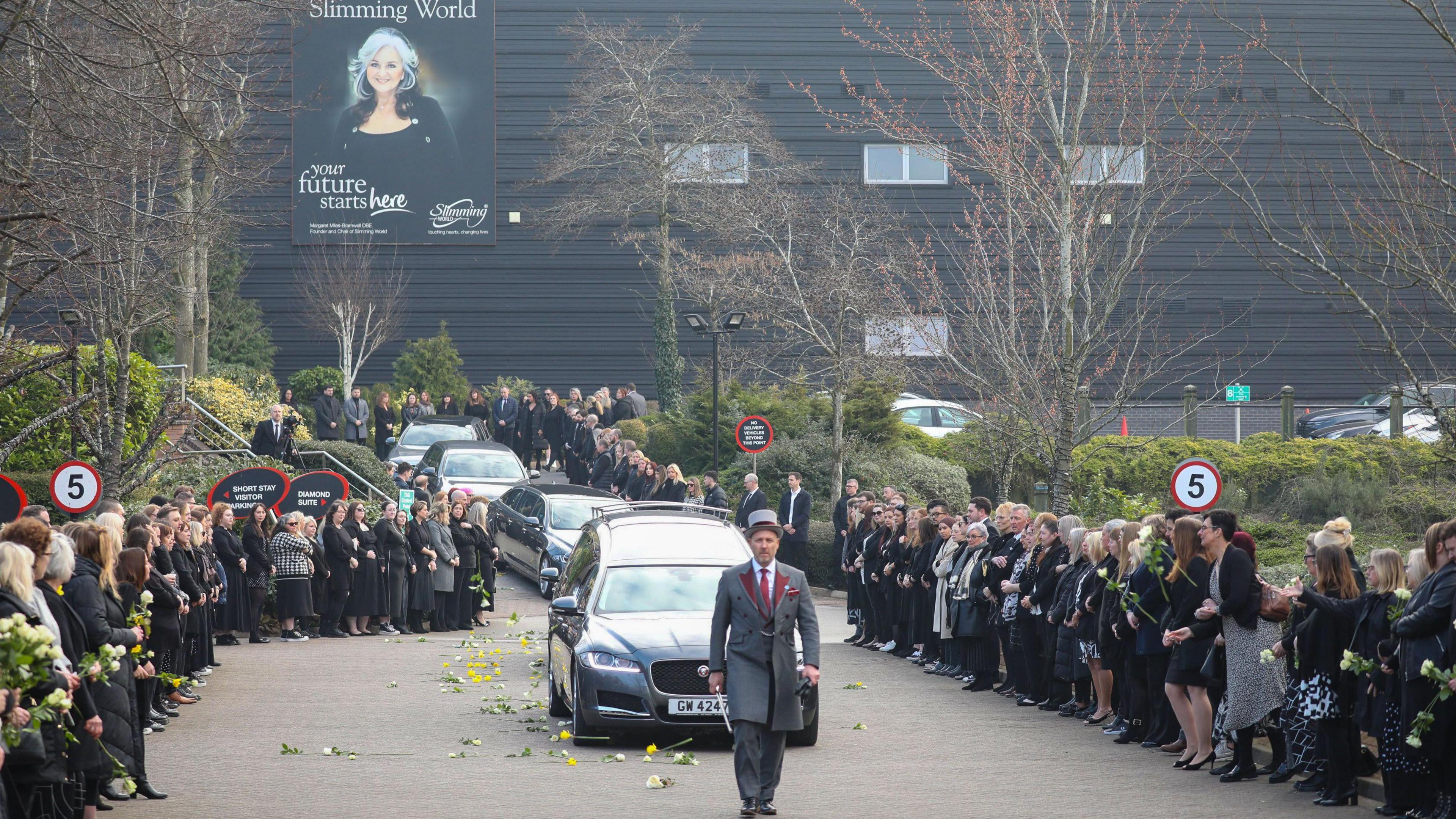 Funeral cortege driving past a large modern office building which has a poster of Margaret Miles-Bramwell on it. Dozens of people line the road on either side.