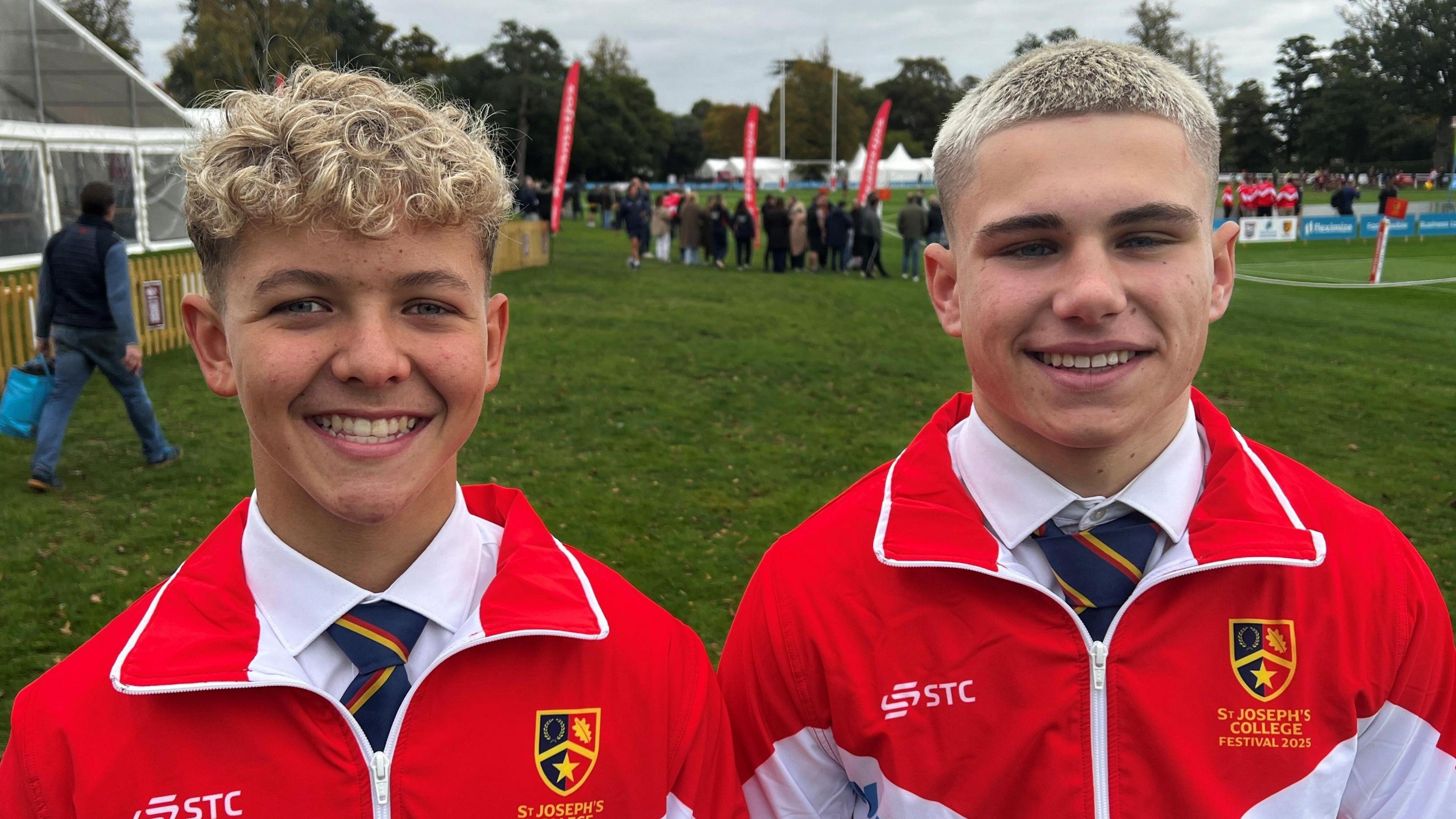 Two boys are standing next to each other, looking at the camera and smiling. They are both wearing red and white tracksuit tops, with white shirts and striped blue ties underneath. They are standing on a playing field.