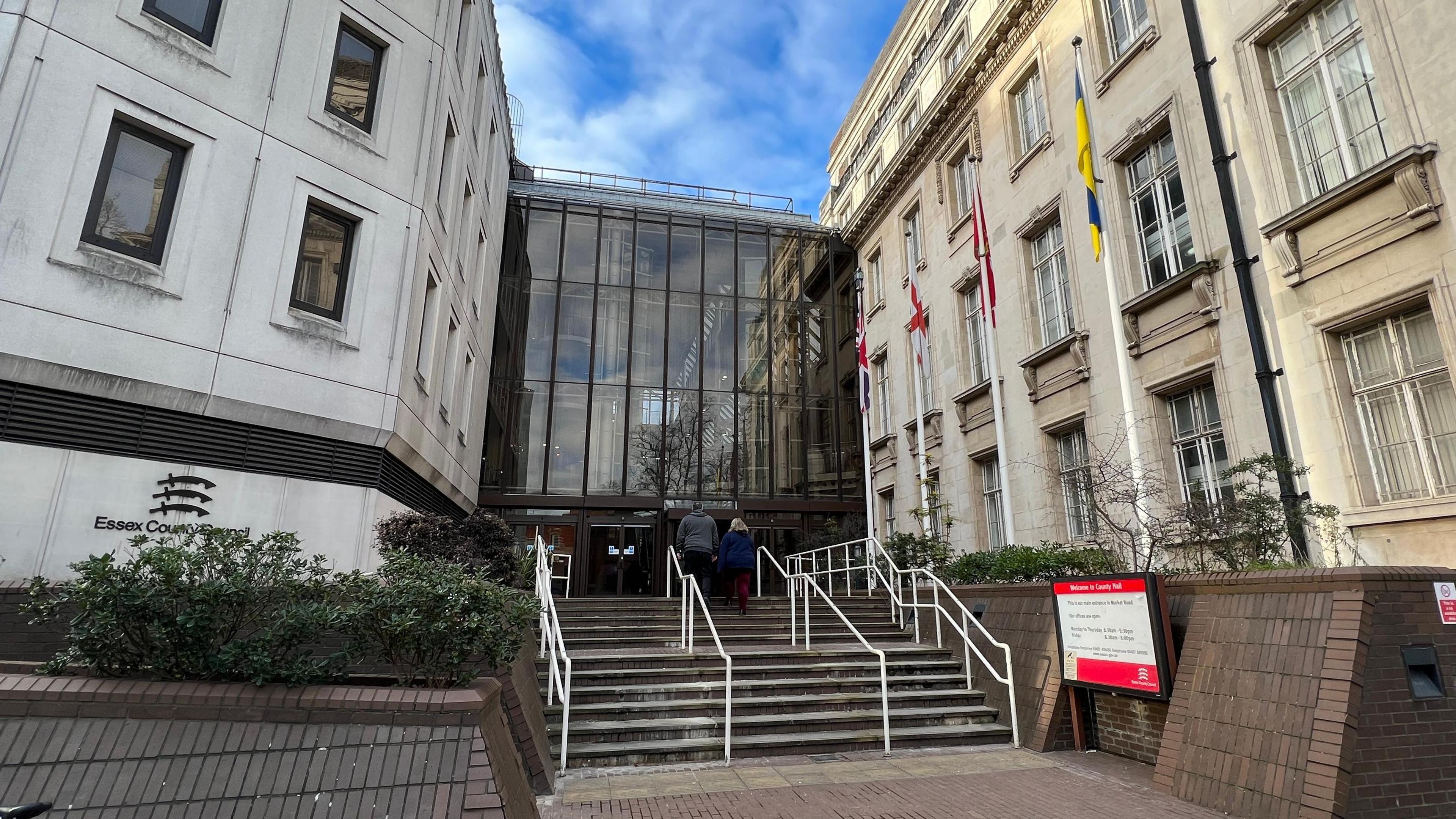 Steps leading to Essex County Council's offices in Chelmsford. The large building is tall and made of cream bricks. It has a tall glass facade in the middle and several flags - including the Union Jack and St George's Cross - are flown on poles outside.