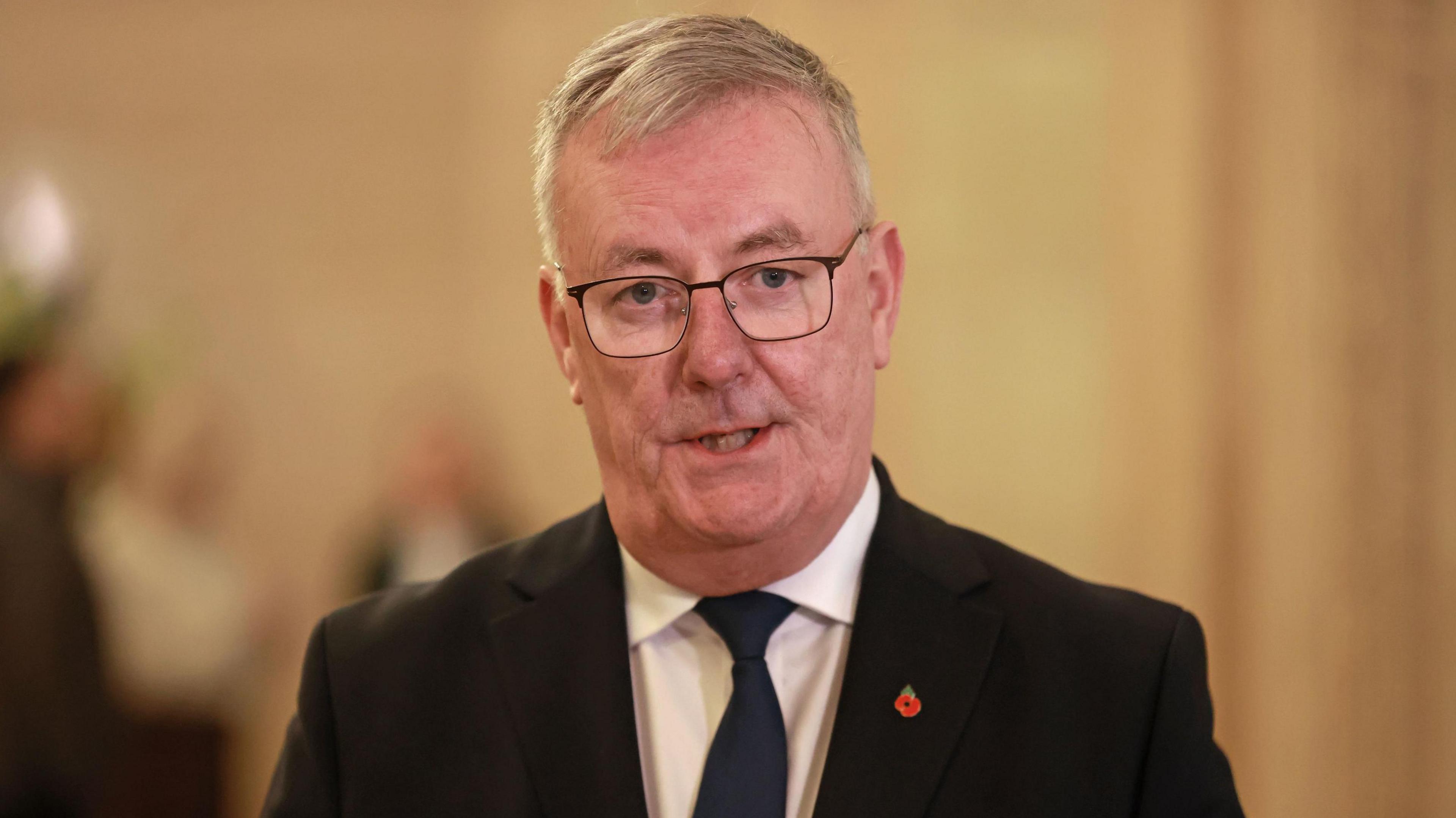 Health Minister Mike Nesbitt wearing a dark suit with a poppy badge on the lapel. There is a blurred background. 