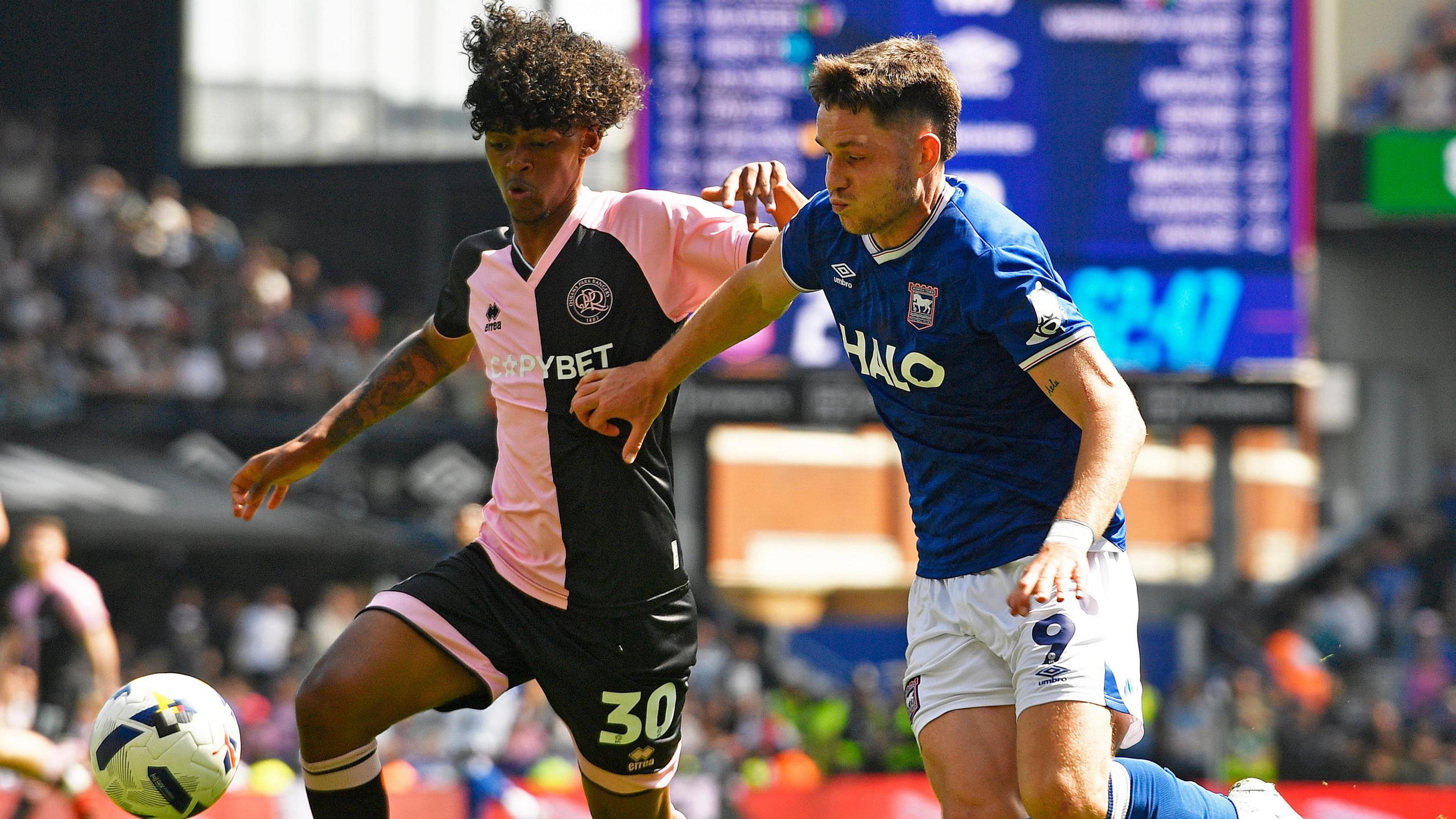 Tylon Smith in pink and navy halved shirt and navy shorts, competes for the ball with an Ipswich player in blue shirt and white shorts