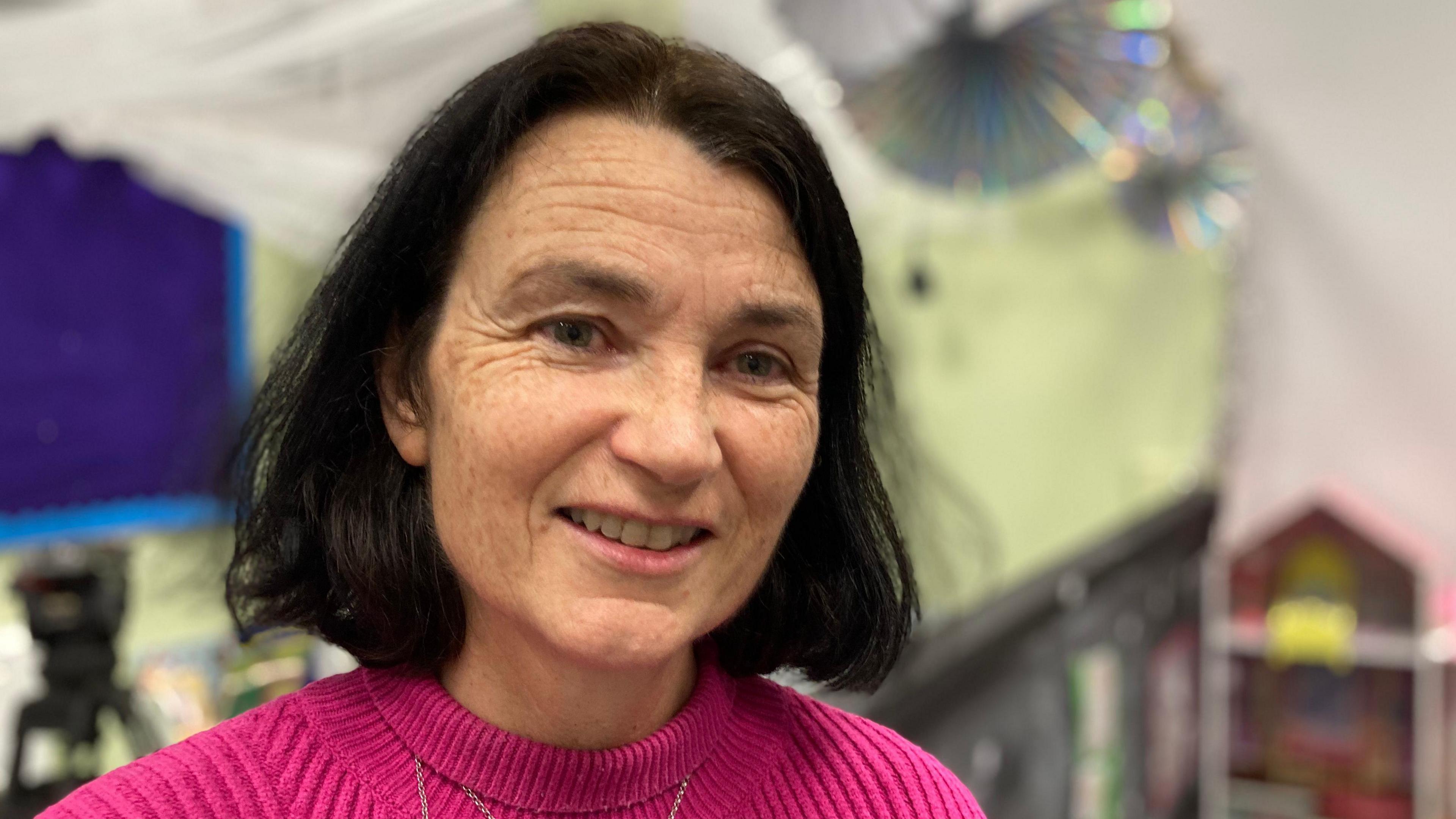 Joan Henderson with shoulder length black hair is smiling at the camera as she wears a pink top.