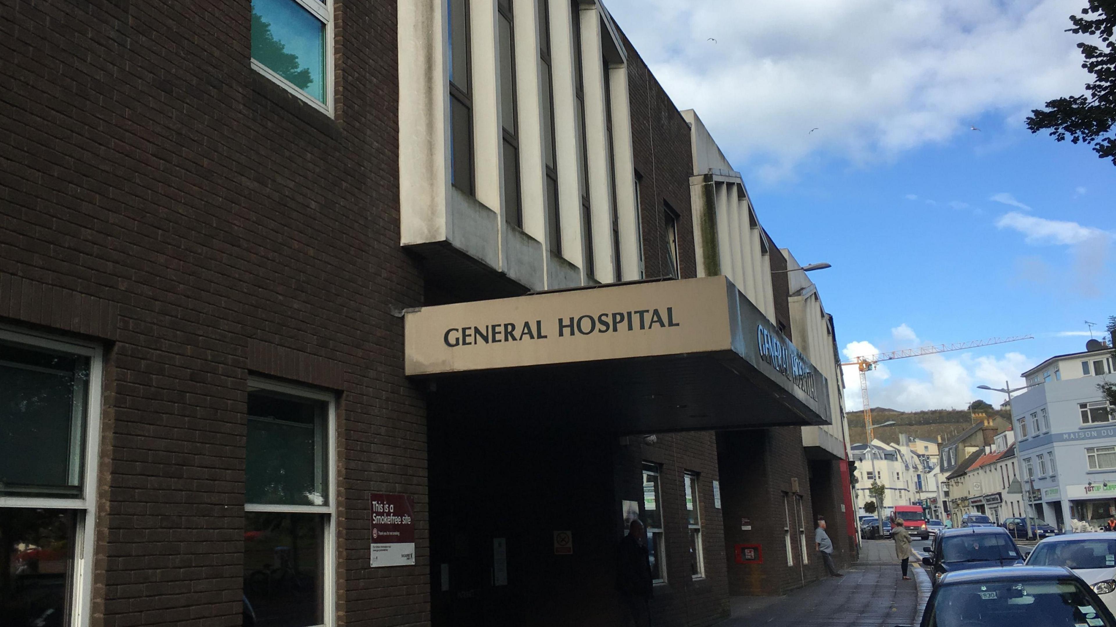 General hospital in Jersey - building on the left with GENERAL HOSPITAL sign above the entrance, road to the right with cars parked along the side, cloudy blue skies.