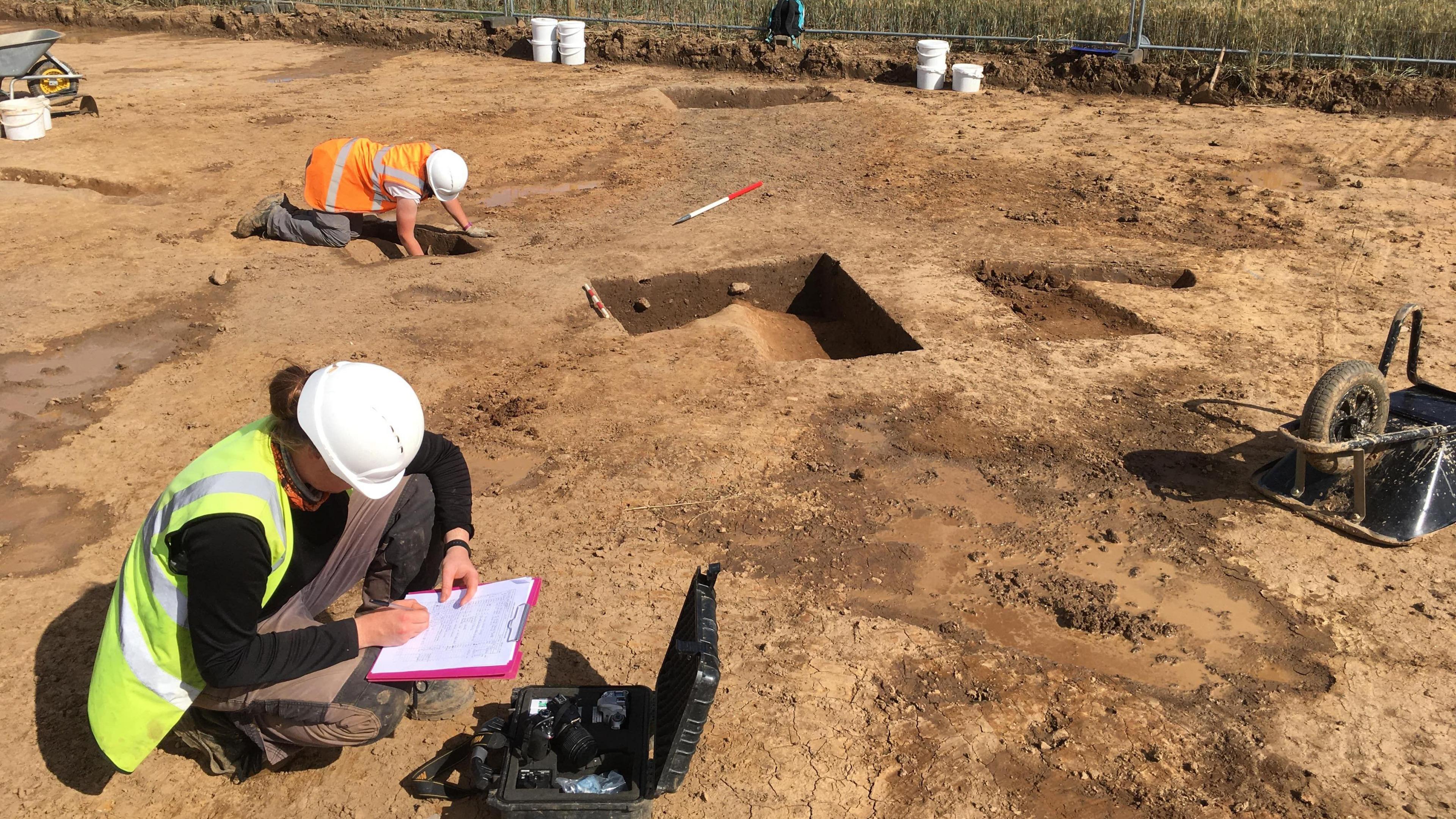 People wearing hard hats and hi-vis vests look into trenches and make notes on clip-boards at a dig site, the ground is bare earth and you can see wheelbarrows and buckets at the scene.