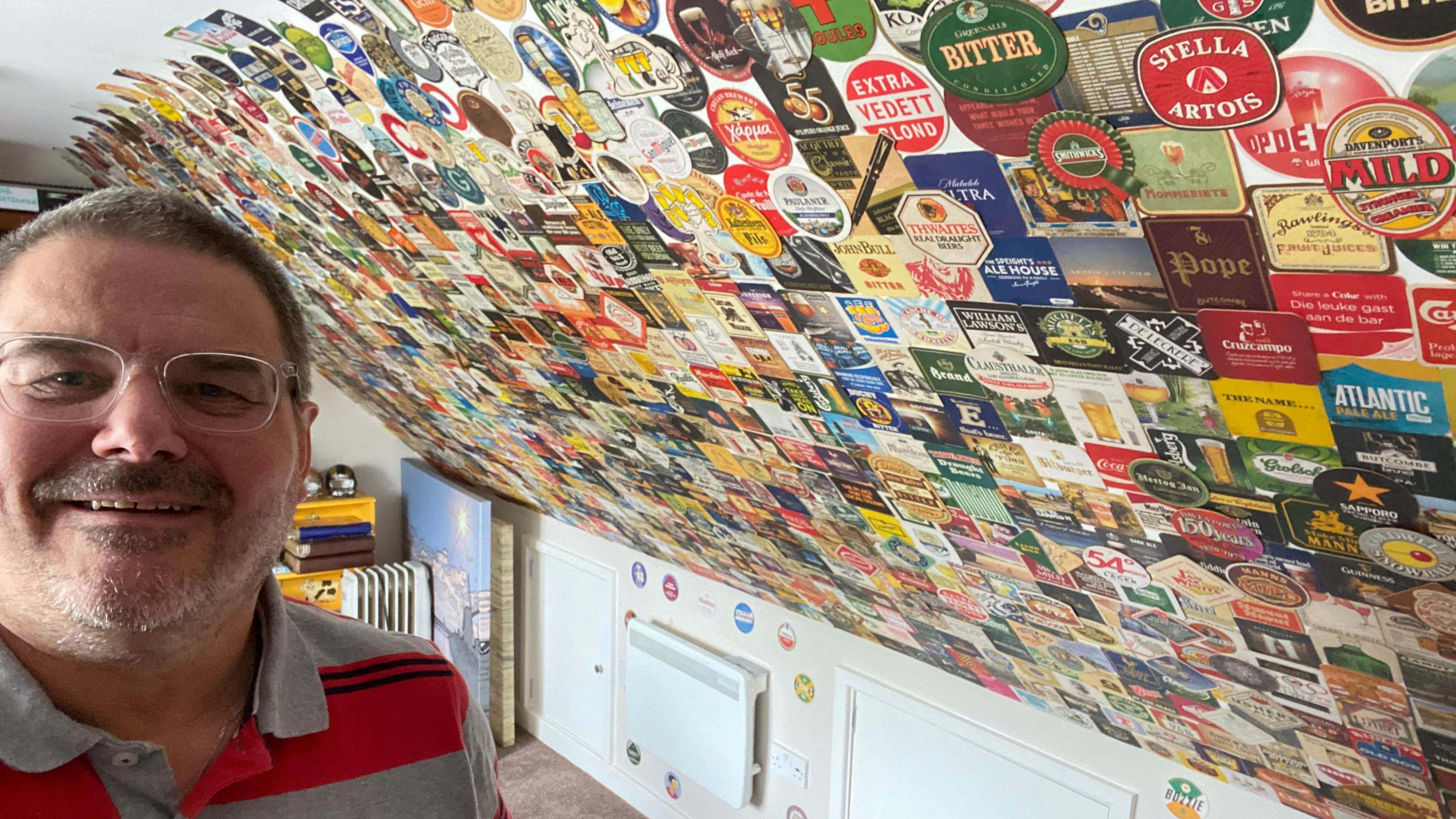 Selfie of a man in the bottom left of the frame, bespectacled and smiling with a wall covered with beer mats next to him