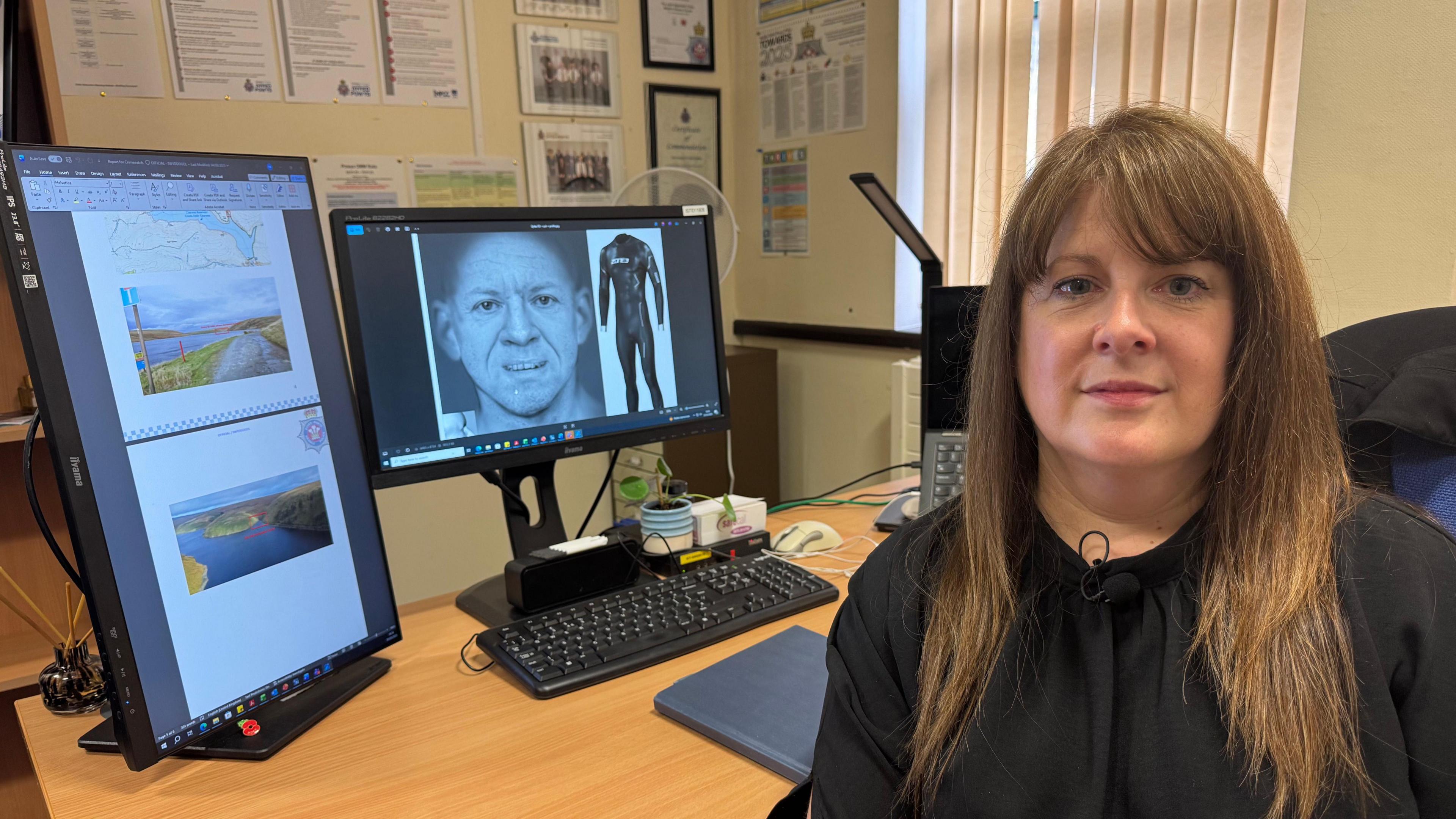 Dynes gyda gwallt hir brown. Mae hi'n gwisgo blows ddu. Mae hi mewn swyddfa, ac yn eistedd o flaen desg. Mae dwy sgrin cyfrifiadur y tu ôl iddi, gyda lluniau digidol o wyneb y dyn, siwt wlyb a lluniau o'r ardal ble y cafodd y dyn ei ddarganfod. 